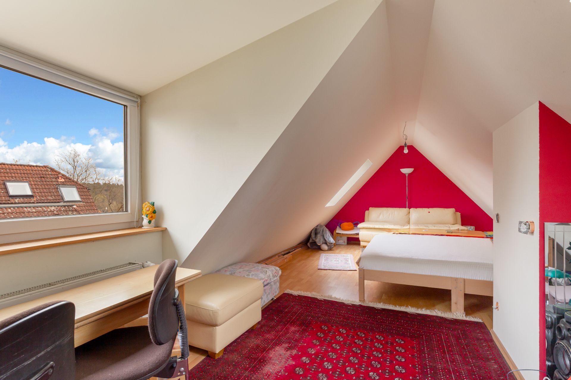 Helles Dachzimmer mit Schreibtisch, Bett, rotem Teppich und Sofa unter Schrägdach.