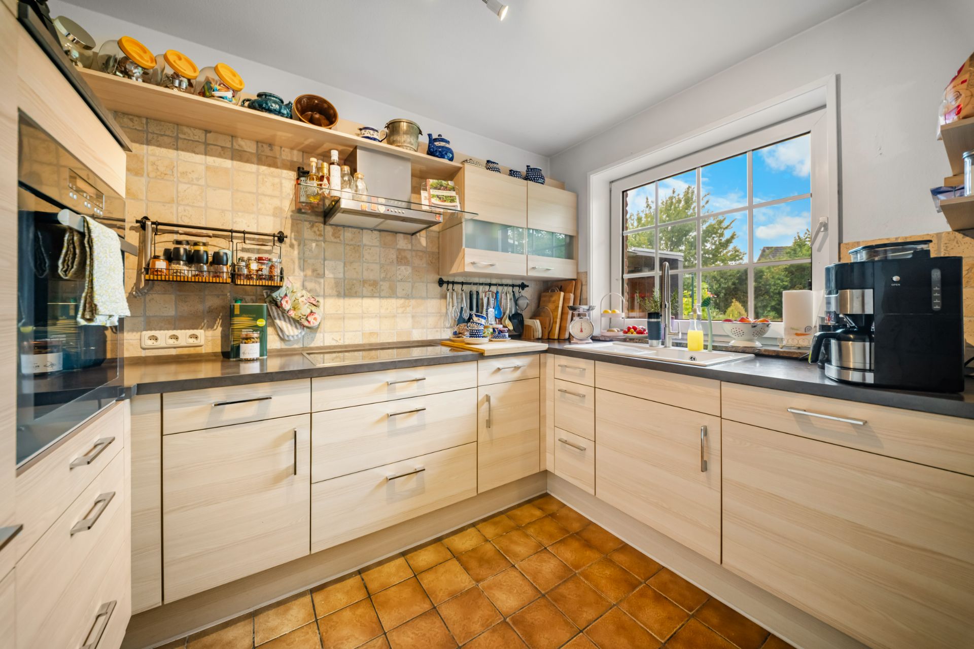 Helle Küche mit Holzmöbeln, großem Fenster, Küchengeräten und Fliesenboden.
