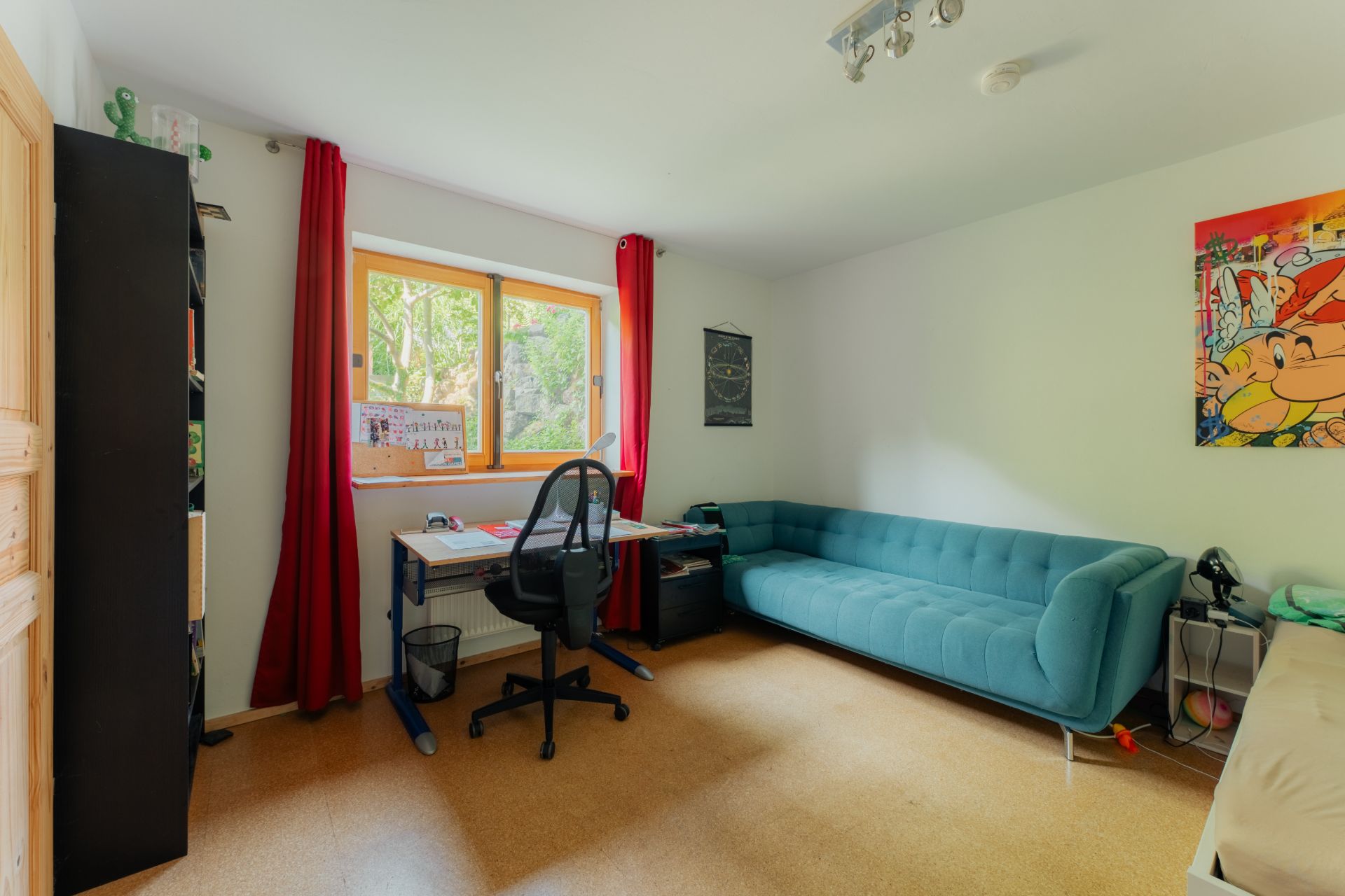 Helles Zimmer mit Schreibtisch, Stuhl, Regal, blauem Sofa und Fenster mit roten Vorhängen.