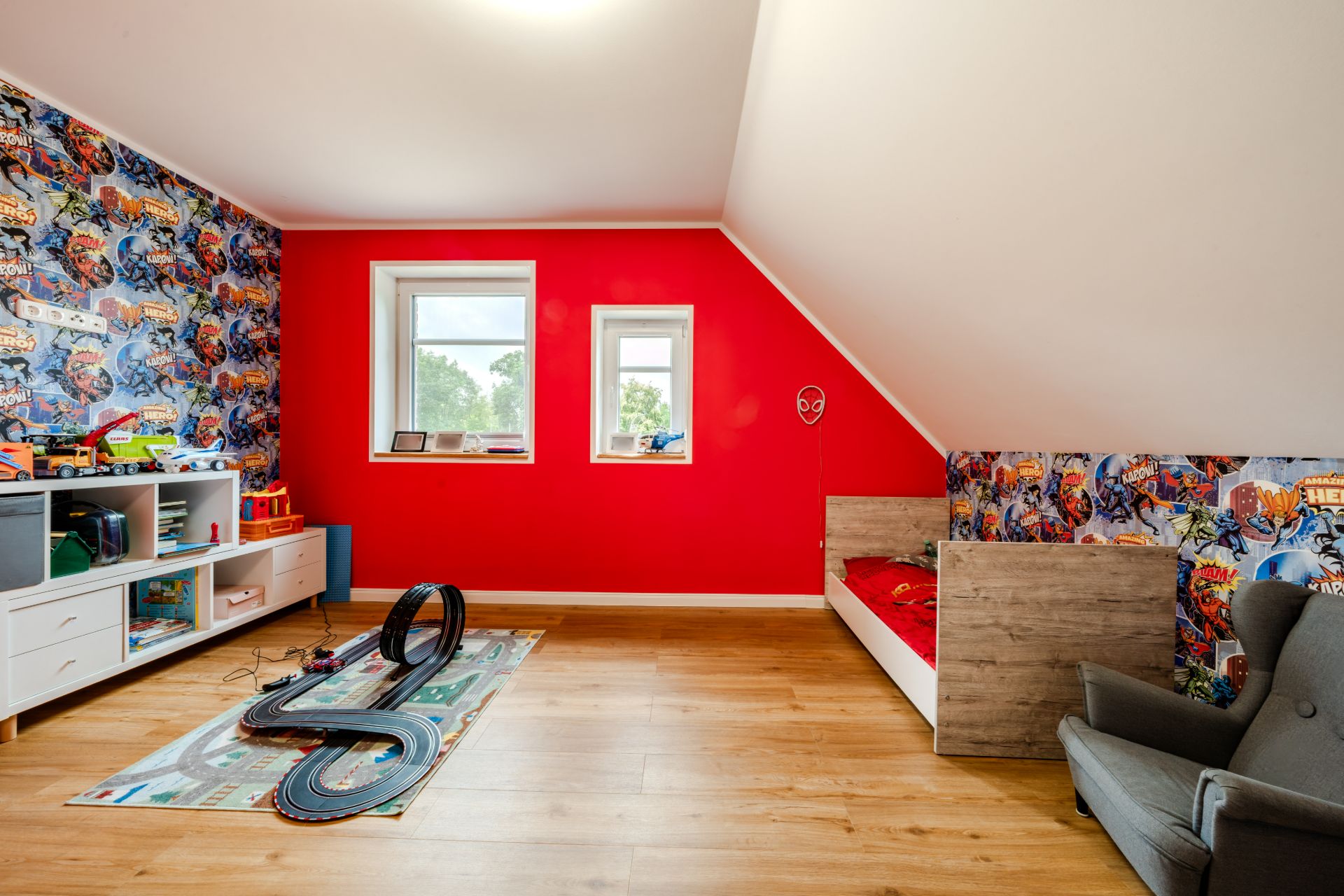 Kinderzimmer mit buntem Comic-Tapeten, roter Wand, Bett, Sessel und Spielzeug-Autorennbahn auf dem Boden.