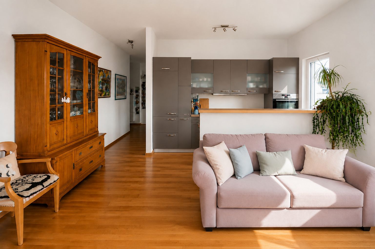Helles Wohnzimmer mit rosa Sofa, Holzboden, großer Vitrine und offener Küche im Hintergrund.