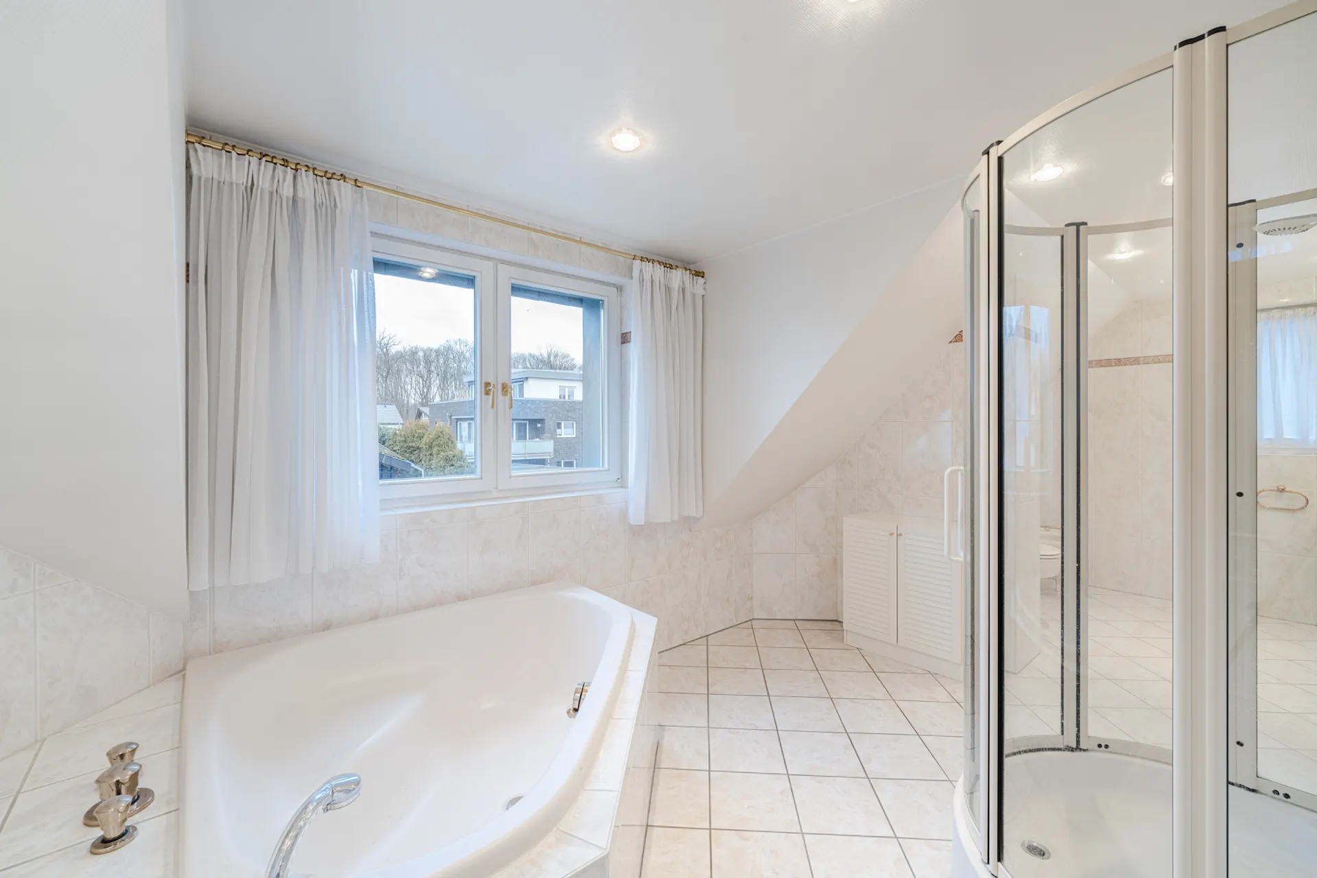 Helles Badezimmer mit Eckbadewanne, Duschecke und Fenster mit Vorhängen.