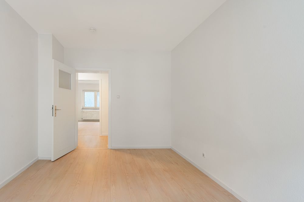 Leerer, heller Raum mit Holzboden, weißer Tür und Fenster im Hintergrund.