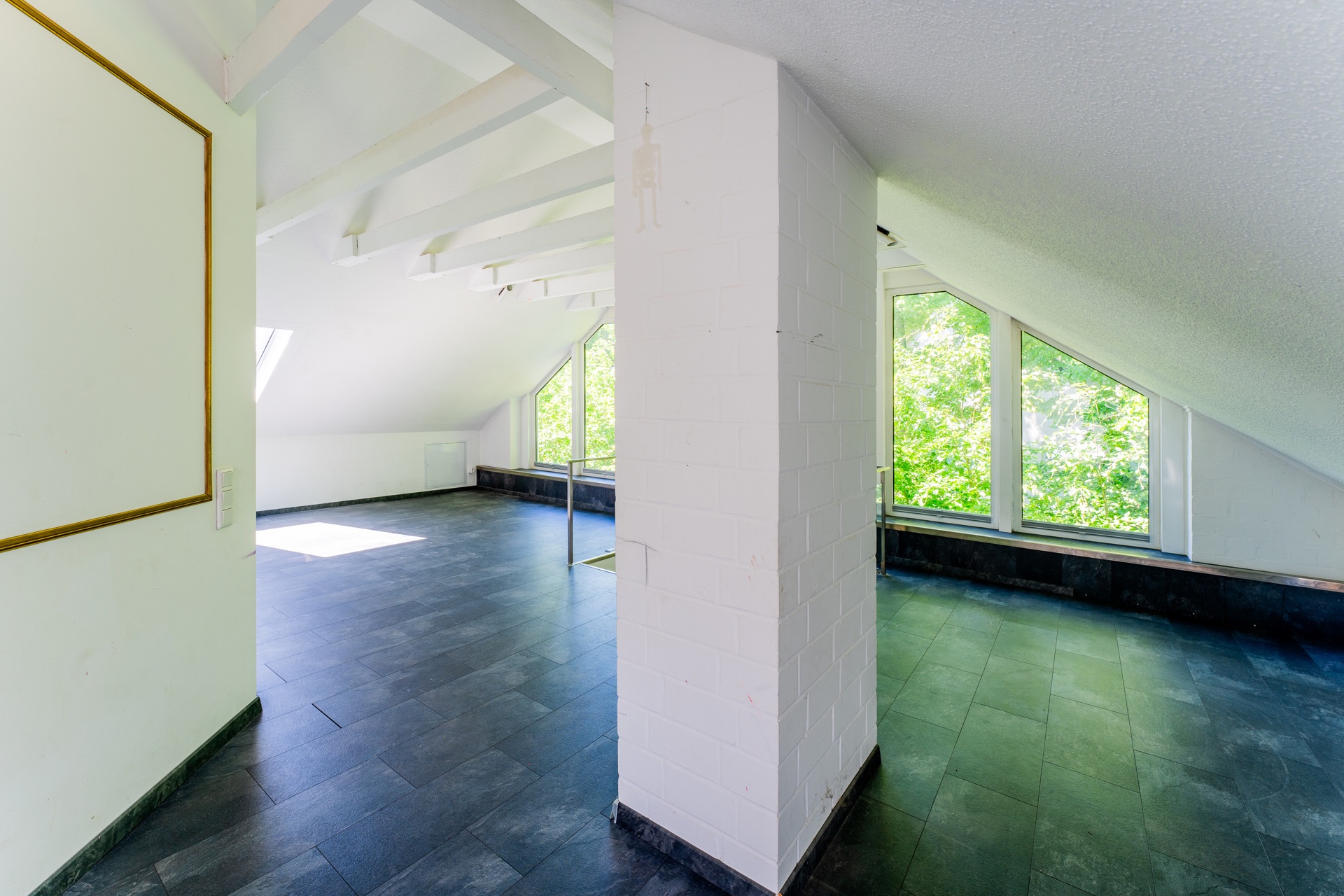 Heller Dachraum mit schräger Decke, weißen Wänden und großen Fenstern mit Blick ins Grüne.