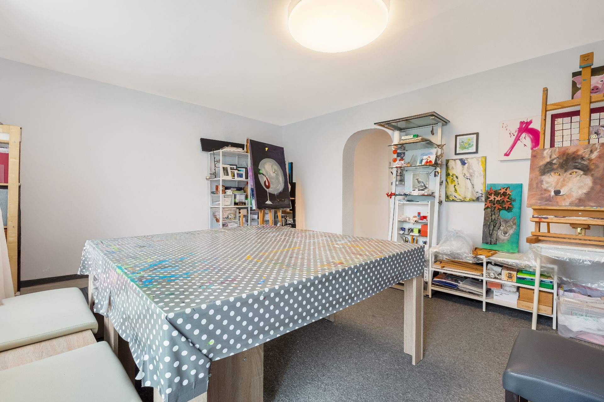 Heller Raum mit großem Tisch, Kunstmaterialien und mehreren Gemälden an der Wand.