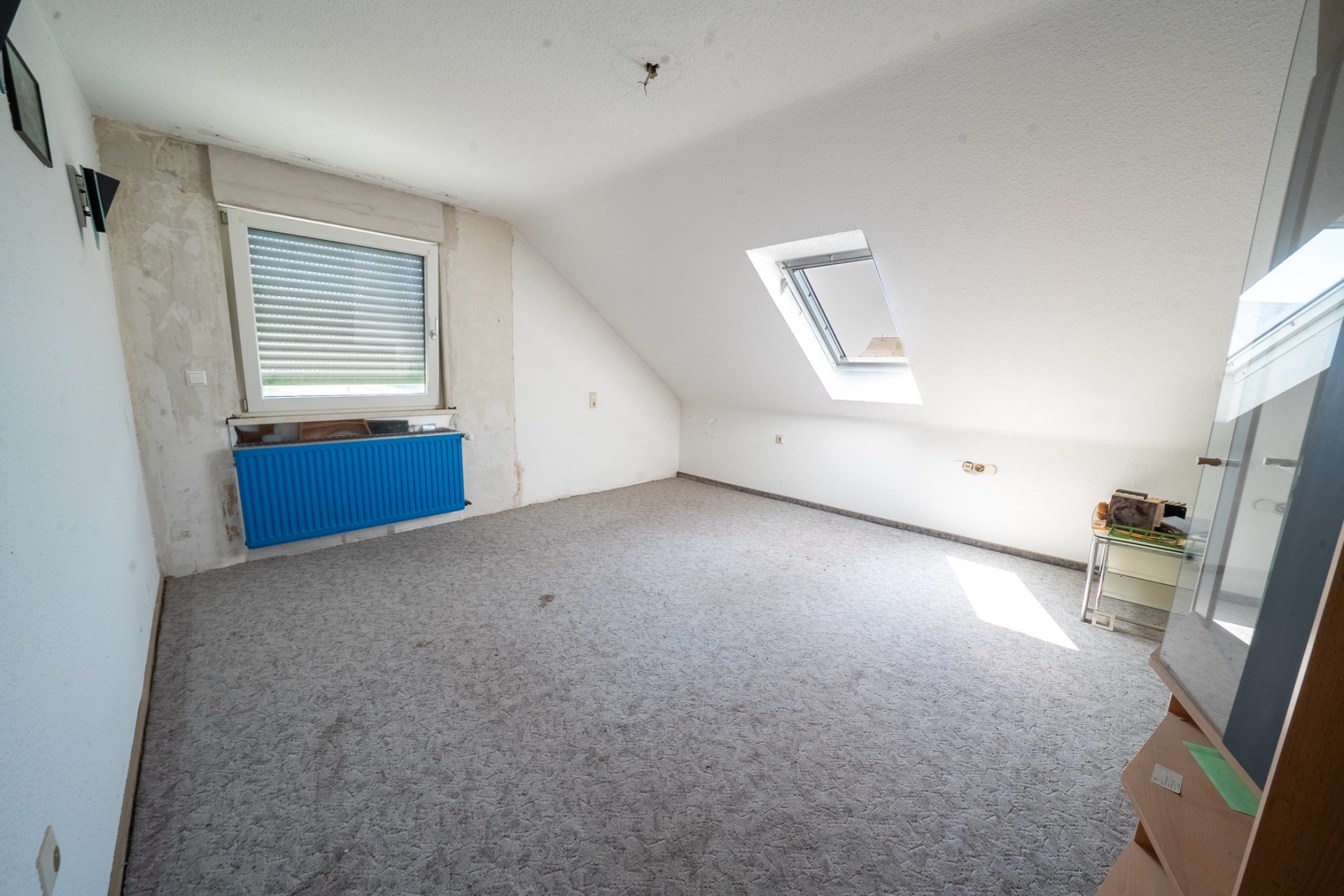 Leeres Zimmer mit Teppichboden, Dachschräge, Dachfenster und blauem Heizkörper.
