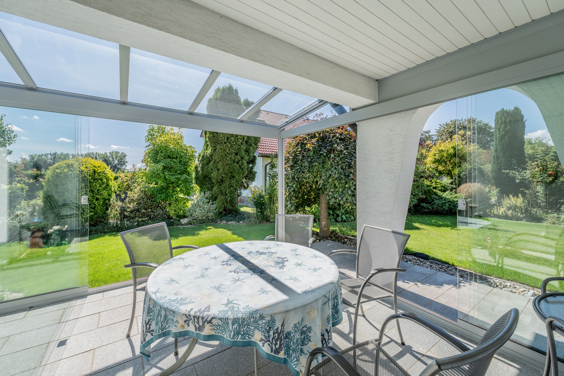 Überdachte Terrasse mit Glastüren, rundem Tisch und Stühlen, Blick auf Garten mit Bäumen und Rasen.