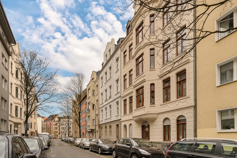 Wohnstraße mit parkenden Autos und mehrstöckigen Wohnhäusern, Bäume ohne Laub.