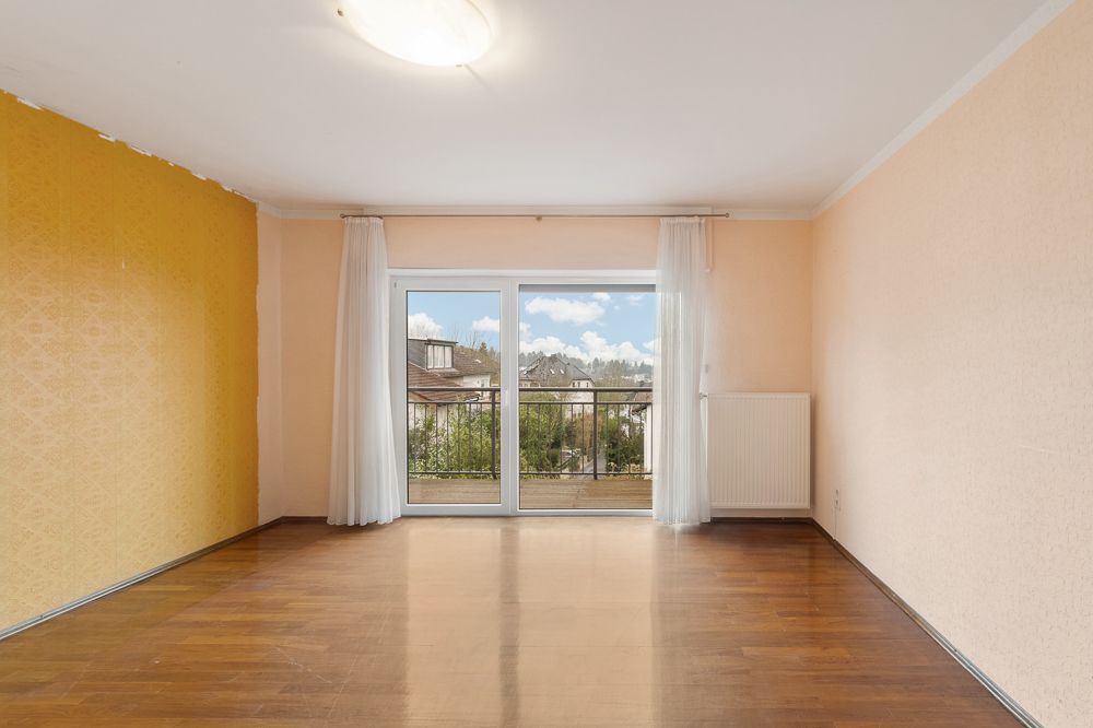 Leeres Zimmer mit Holzfußboden, gelber und beiger Wand sowie Balkon mit Glastür und Vorhängen.