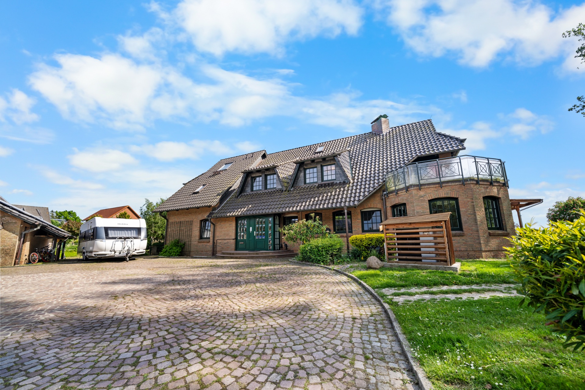 Großes Backsteinhaus mit dunklem Dach, gepflastertem Hof und Wohnwagen auf der linken Seite.