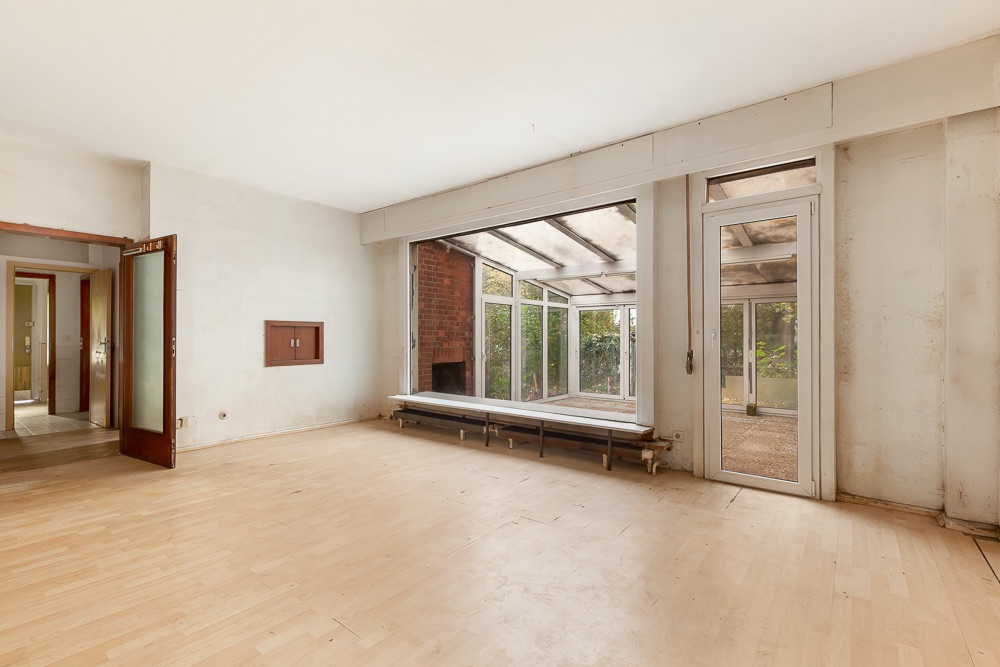 Leeres Wohnzimmer mit Holzboden, großen Fenstern und Zugang zu einem Wintergarten.