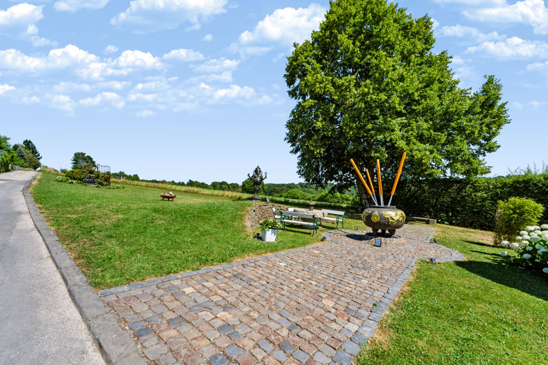 Gepflasterter Weg mit Sitzbänken und Baum im Garten, moderne Skulptur vorne.