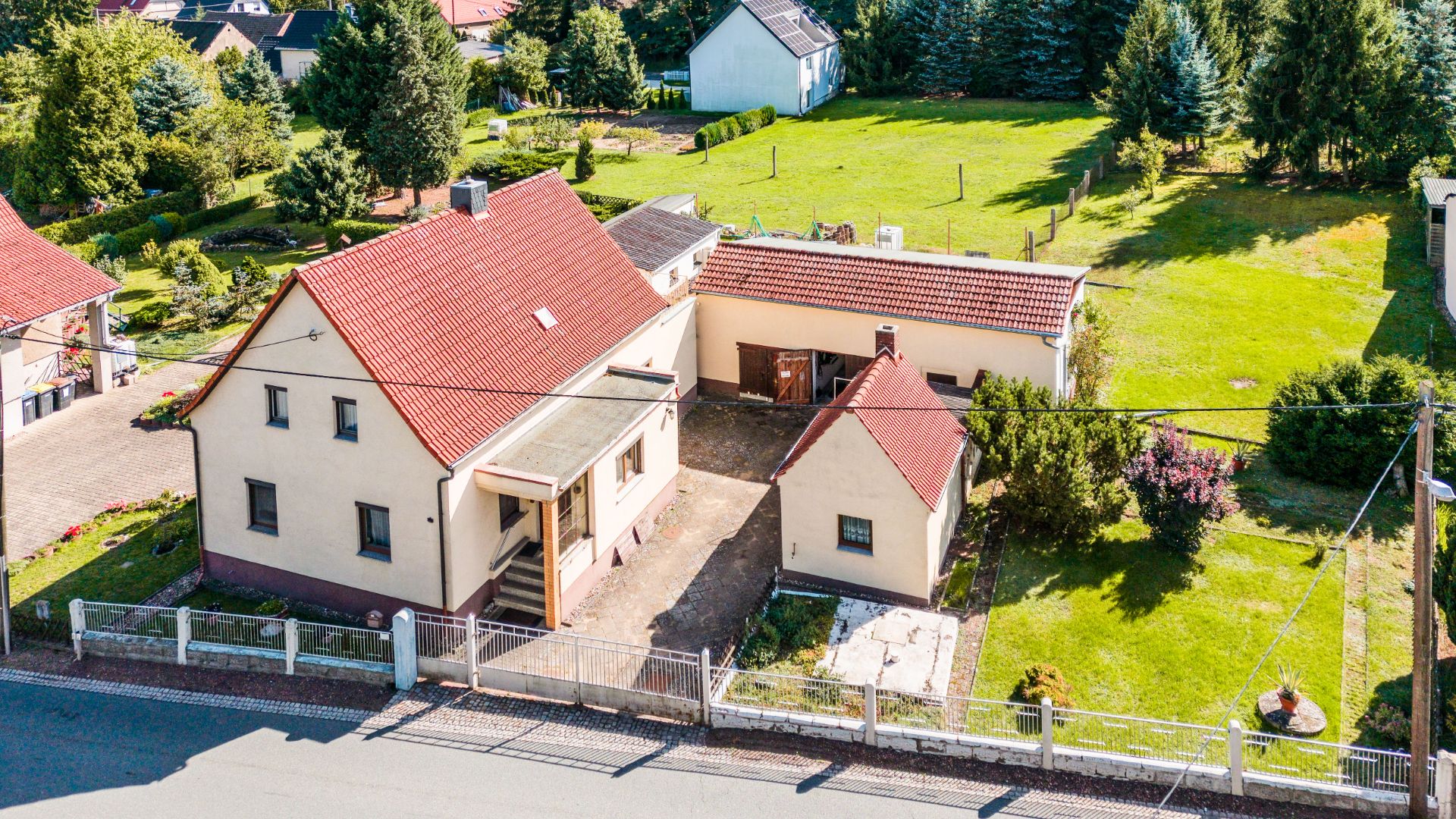 Luftaufnahme eines Wohnhauses mit rotem Dach, Nebengebäude und großem Garten hinter einem Zaun.