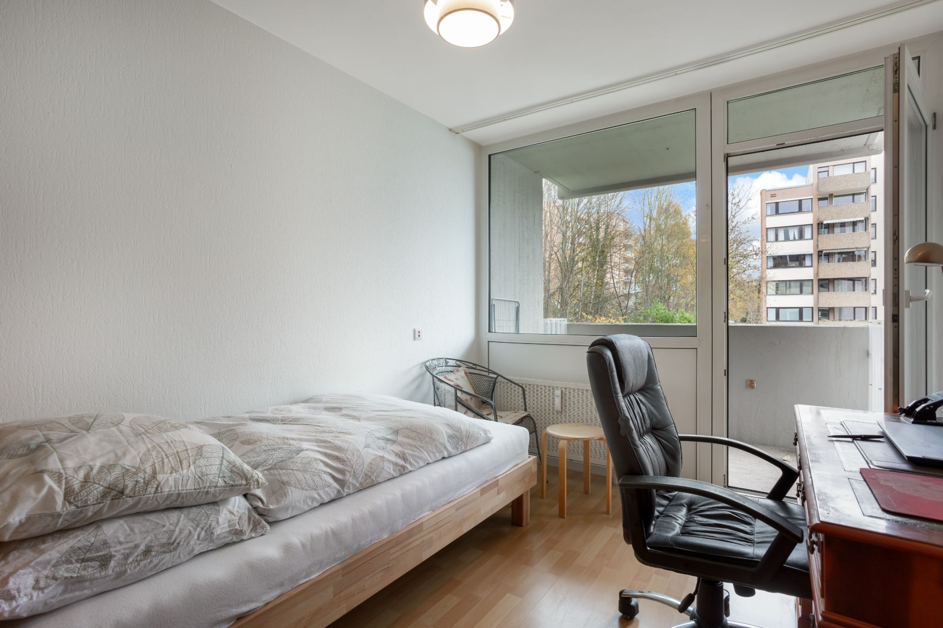 Helles Zimmer mit Einzelbett, Schreibtisch, Bürostuhl und Zugang zu einem Balkon.