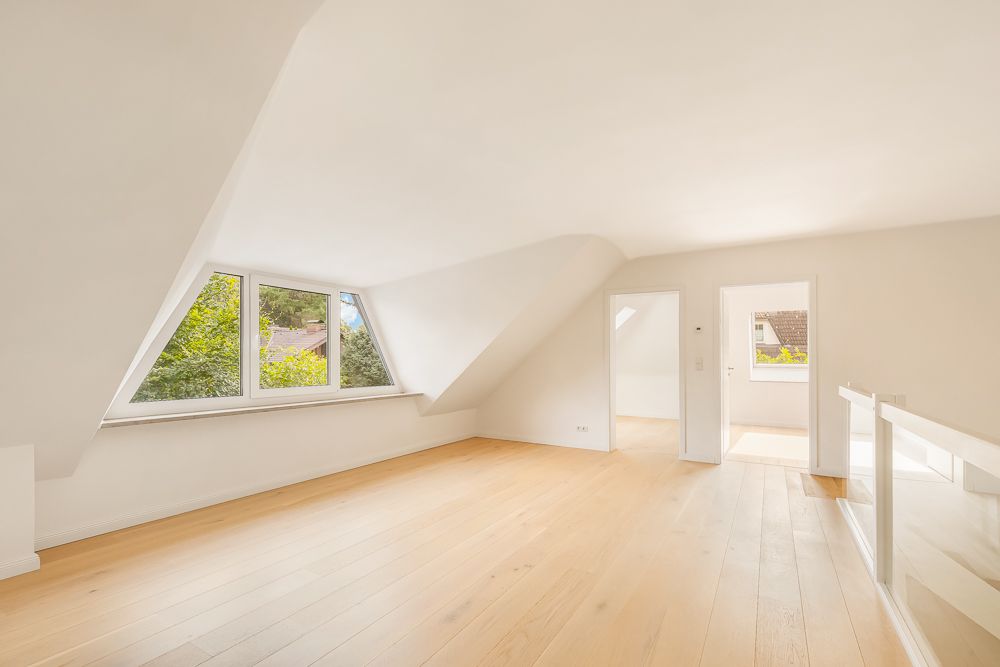 Heller Dachgeschossraum mit großem Fenster, Holzfußboden und weiß gestrichenen Wänden.