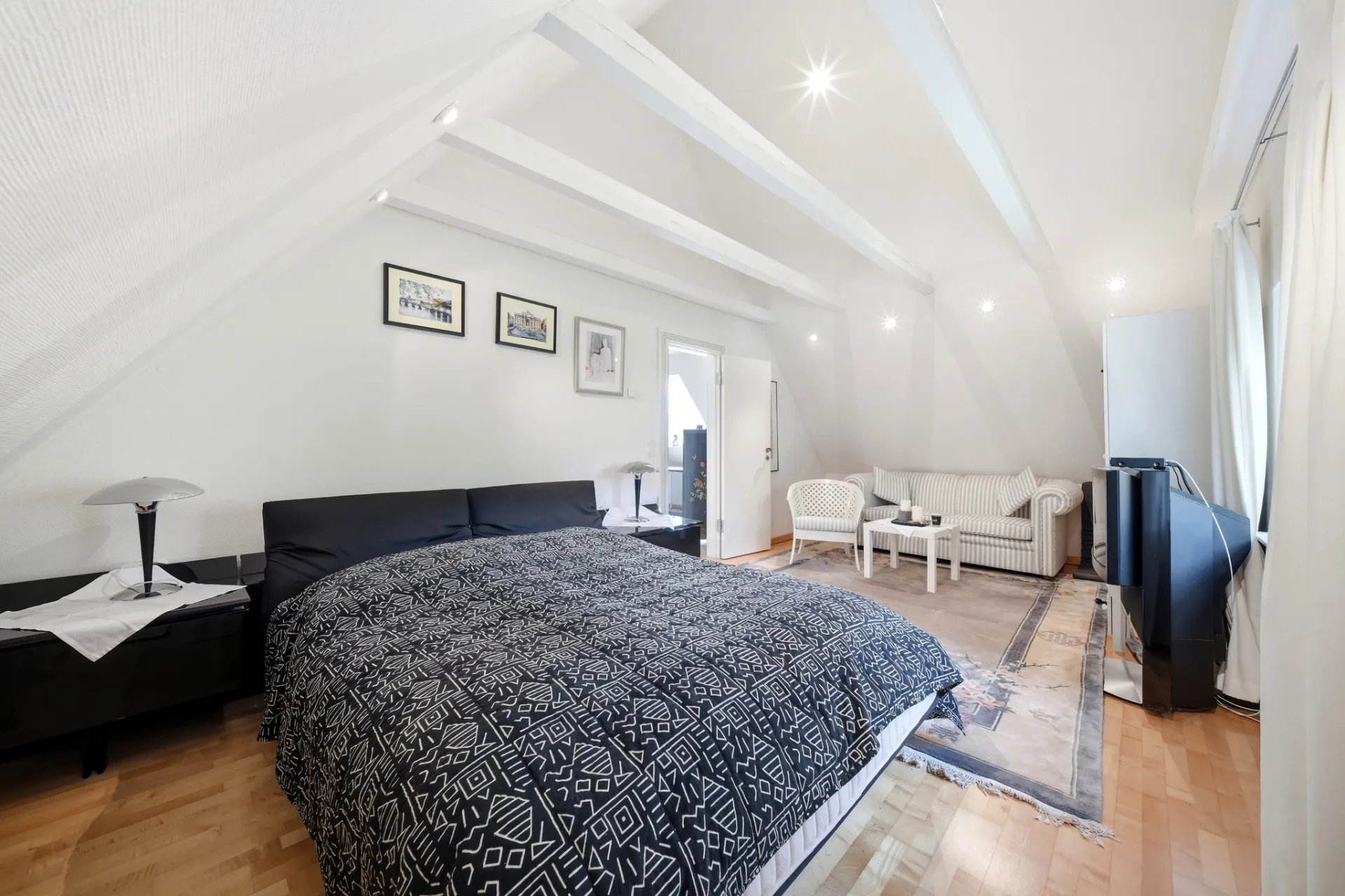 Schlafzimmer mit Doppelbett, Sofa, zwei Sesseln, Fernseher und Bilder an der Wand unter Dachschräge.
