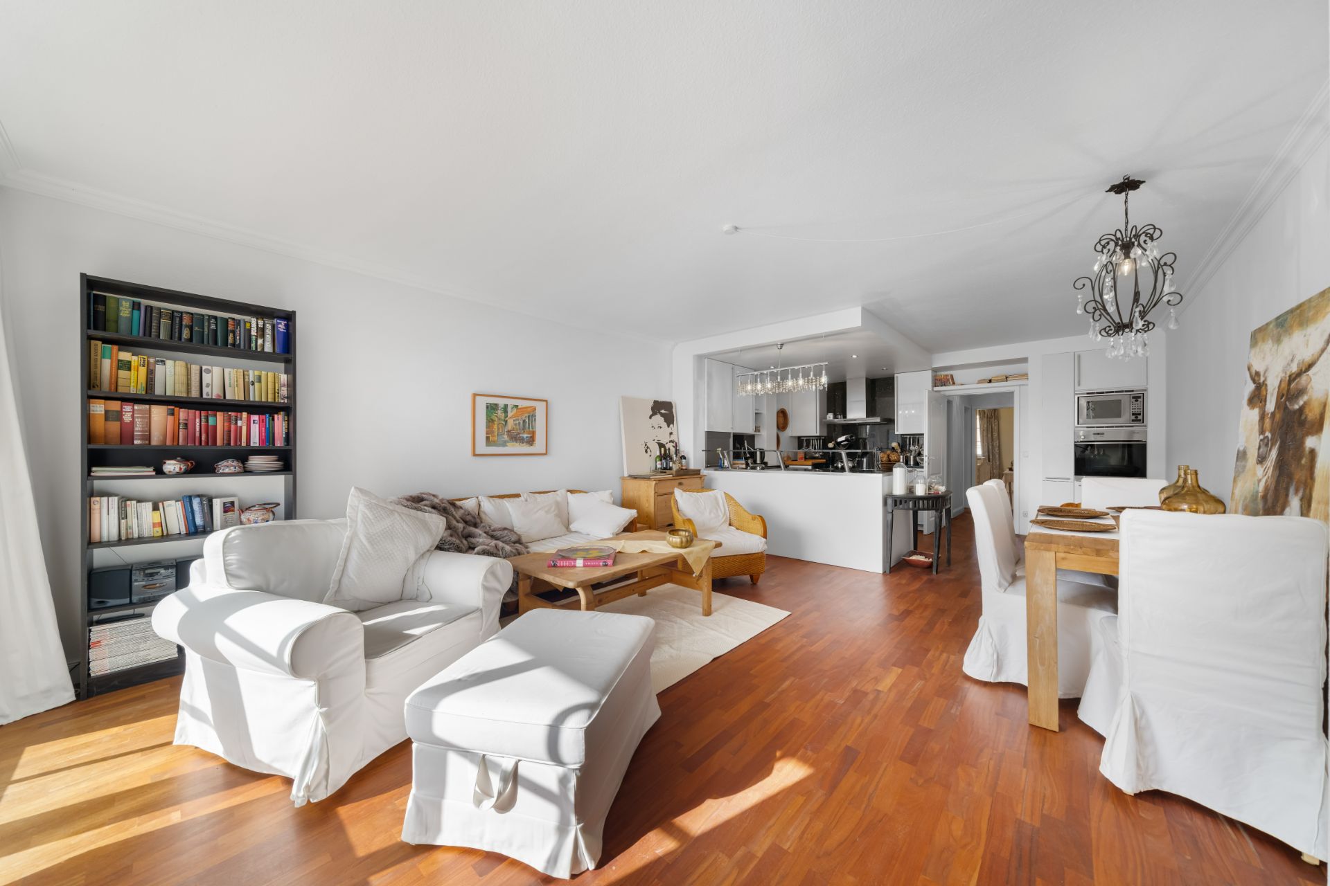 Helles Wohnzimmer mit weißem Sofa, Bücherregal, Esstisch und offener Küche auf Holzfußboden.