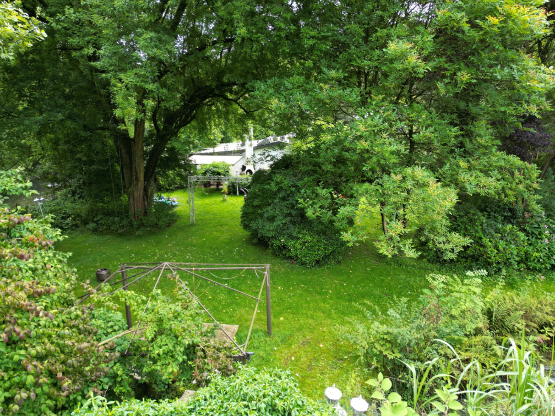 Grüner Garten mit Rasen, Bäumen und Sträuchern, im Hintergrund ein weißes Gebäude.