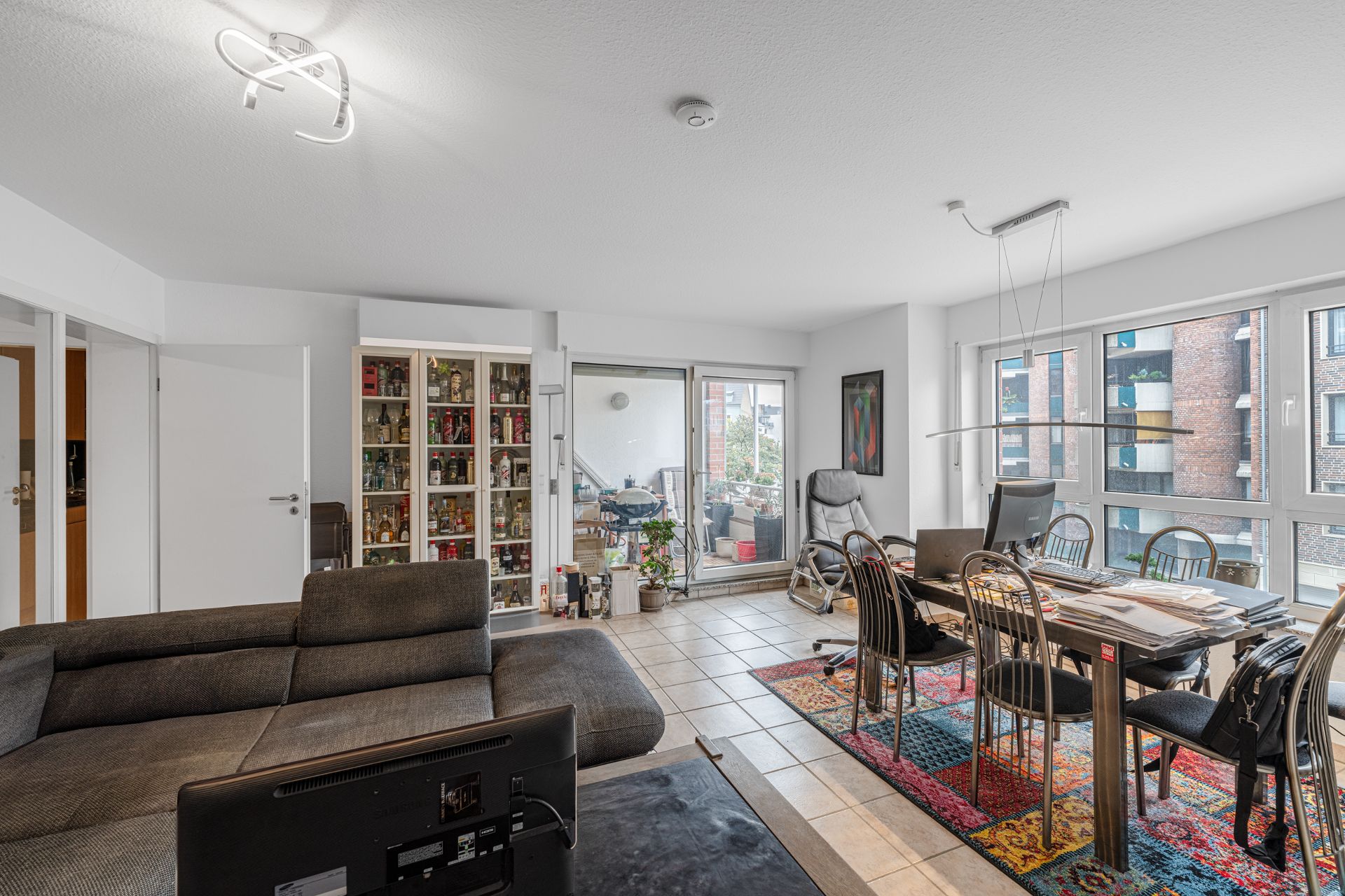 Modernes Wohnzimmer mit Sofa, Esstisch, buntem Teppich und Zugang zum Balkon.