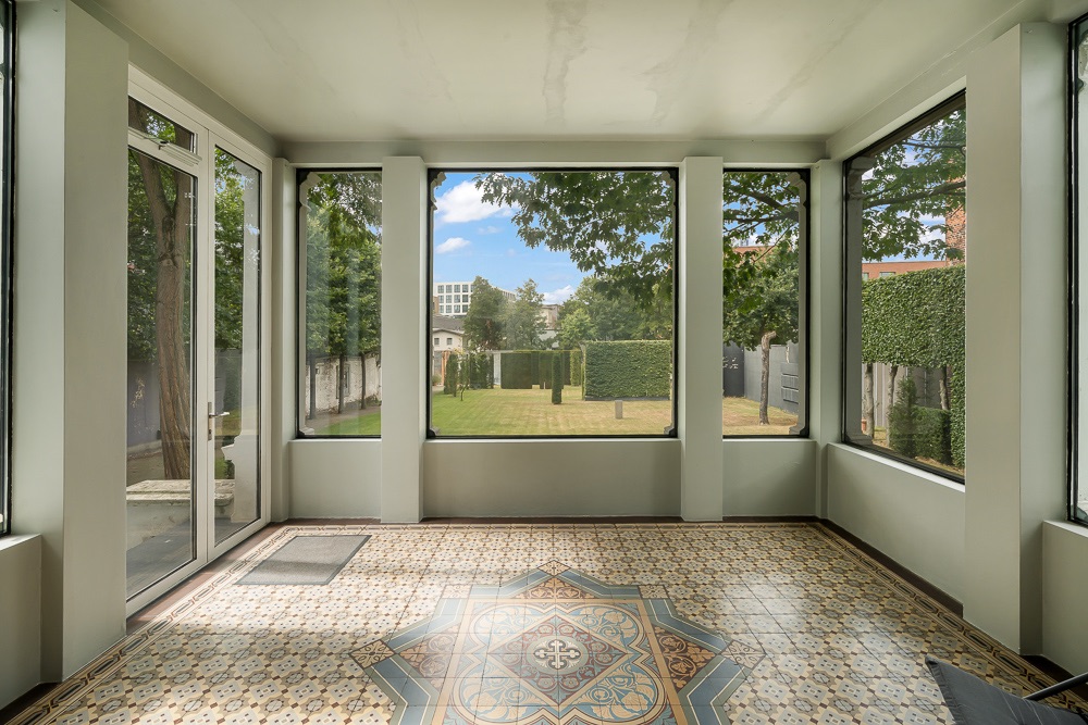 Wintergarten mit großen Fenstern und gemustertem Fliesenboden, Blick auf grünen Garten.