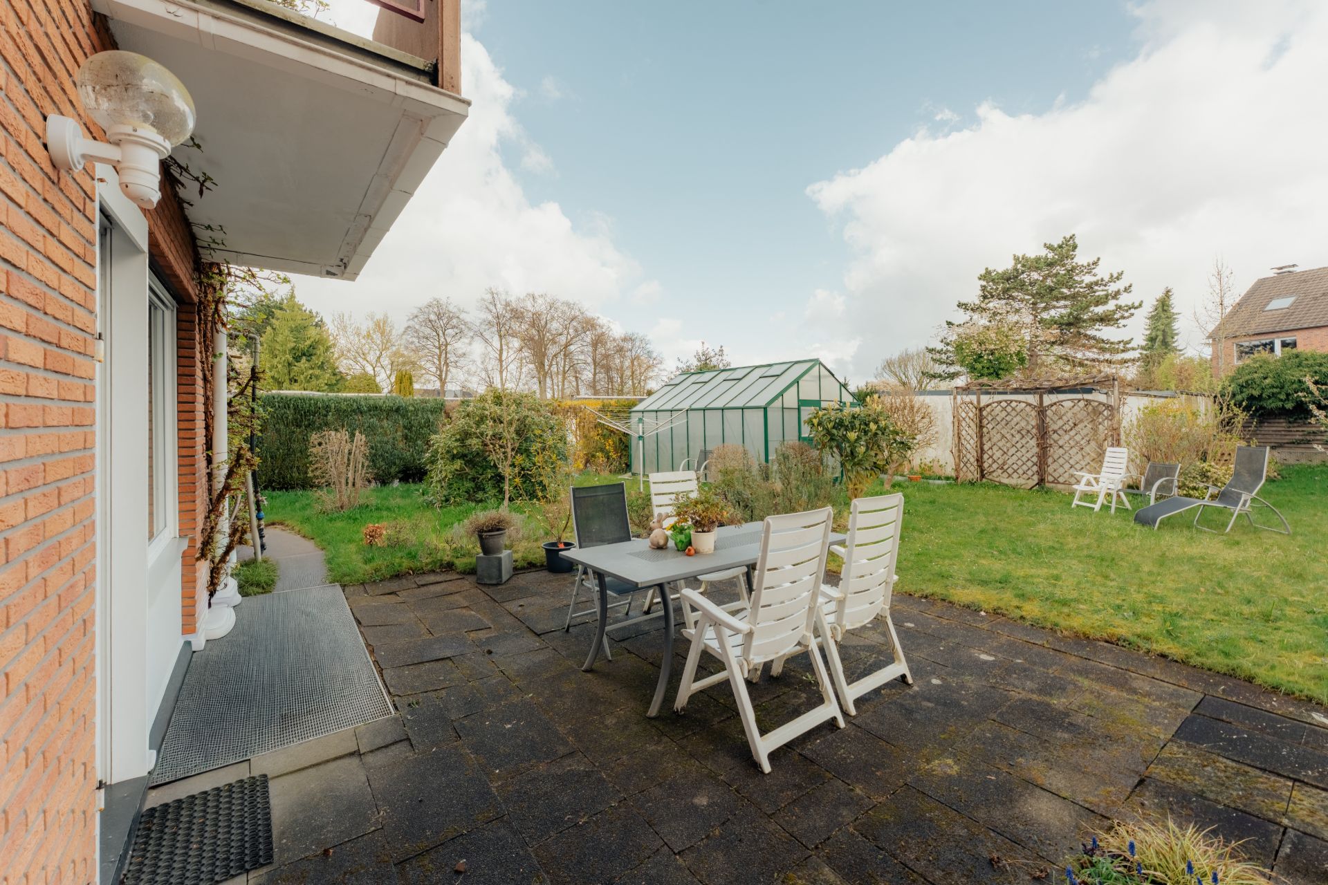 Gepflasterte Terrasse mit Tisch und Stühlen, angrenzender Garten und Gewächshaus im Hintergrund.