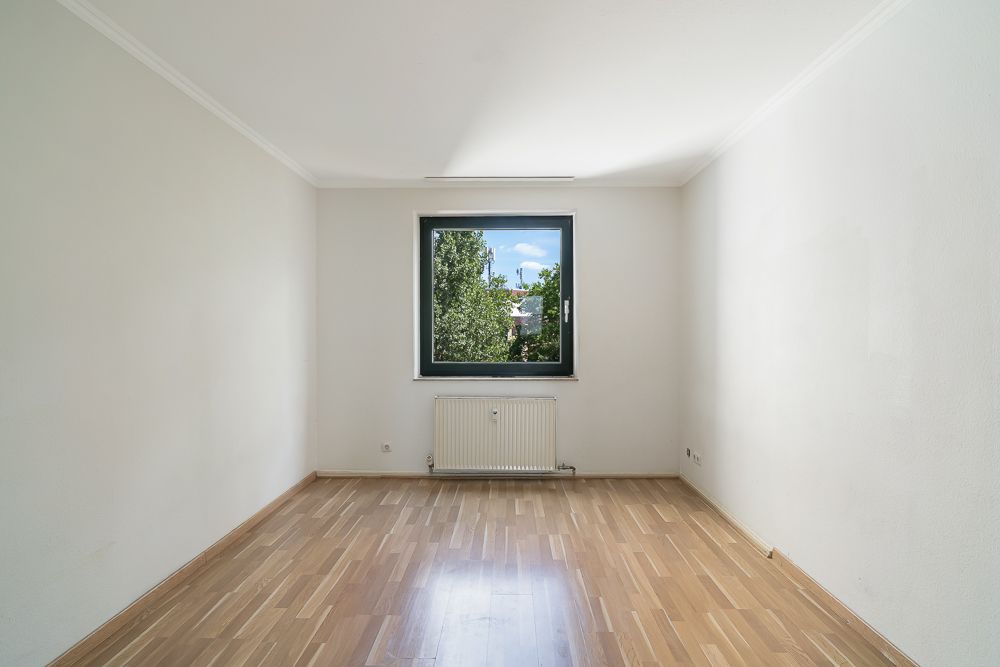 Leeres Zimmer mit Holzfußboden, weiß gestrichenen Wänden und einem Fenster mit Blick ins Grüne.