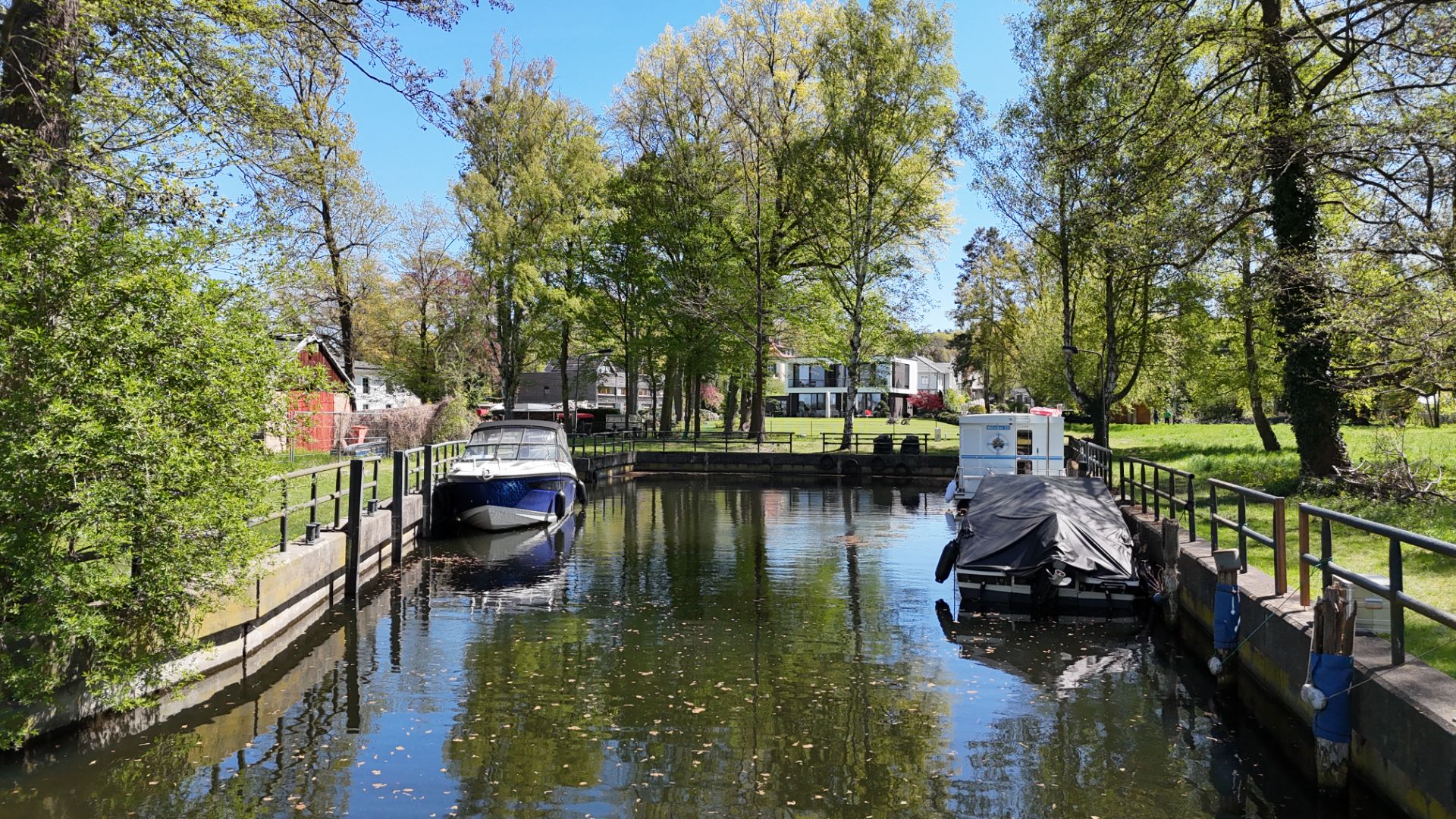 Kleiner Kanal mit zwei Booten, umgeben von Bäumen und Wohnhäusern im Hintergrund.