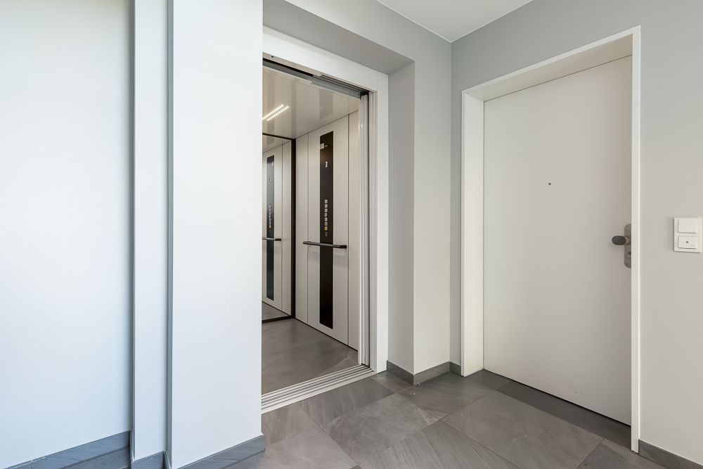 Offener Aufzug und weiße Wohnungstür in einem hellen Flur mit grauem Boden.