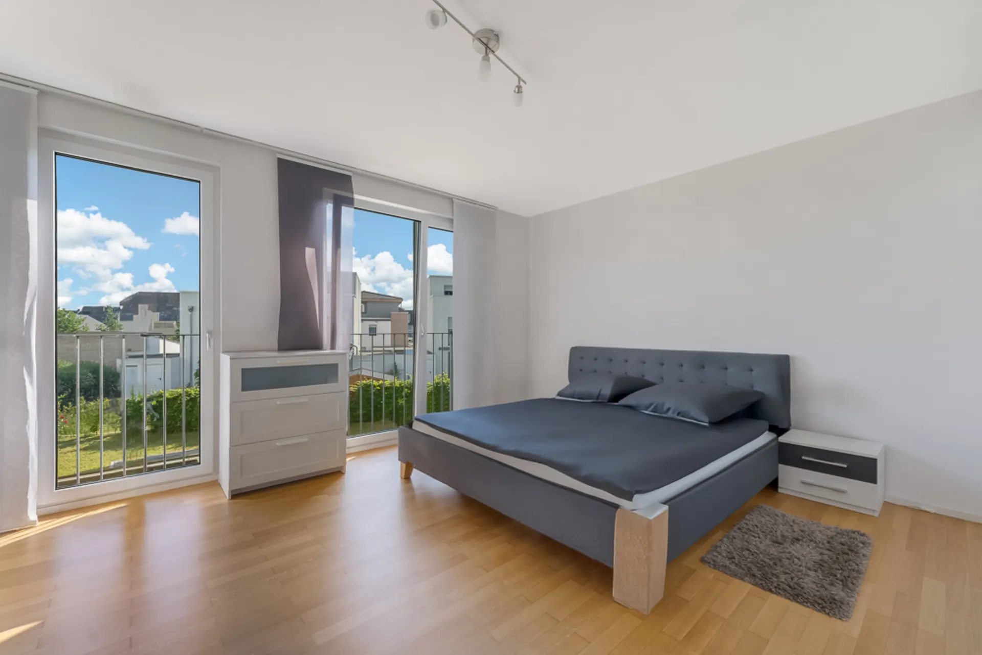 Helles Schlafzimmer mit Doppelbett, Nachttisch, Kommode und zwei großen Fenstern.