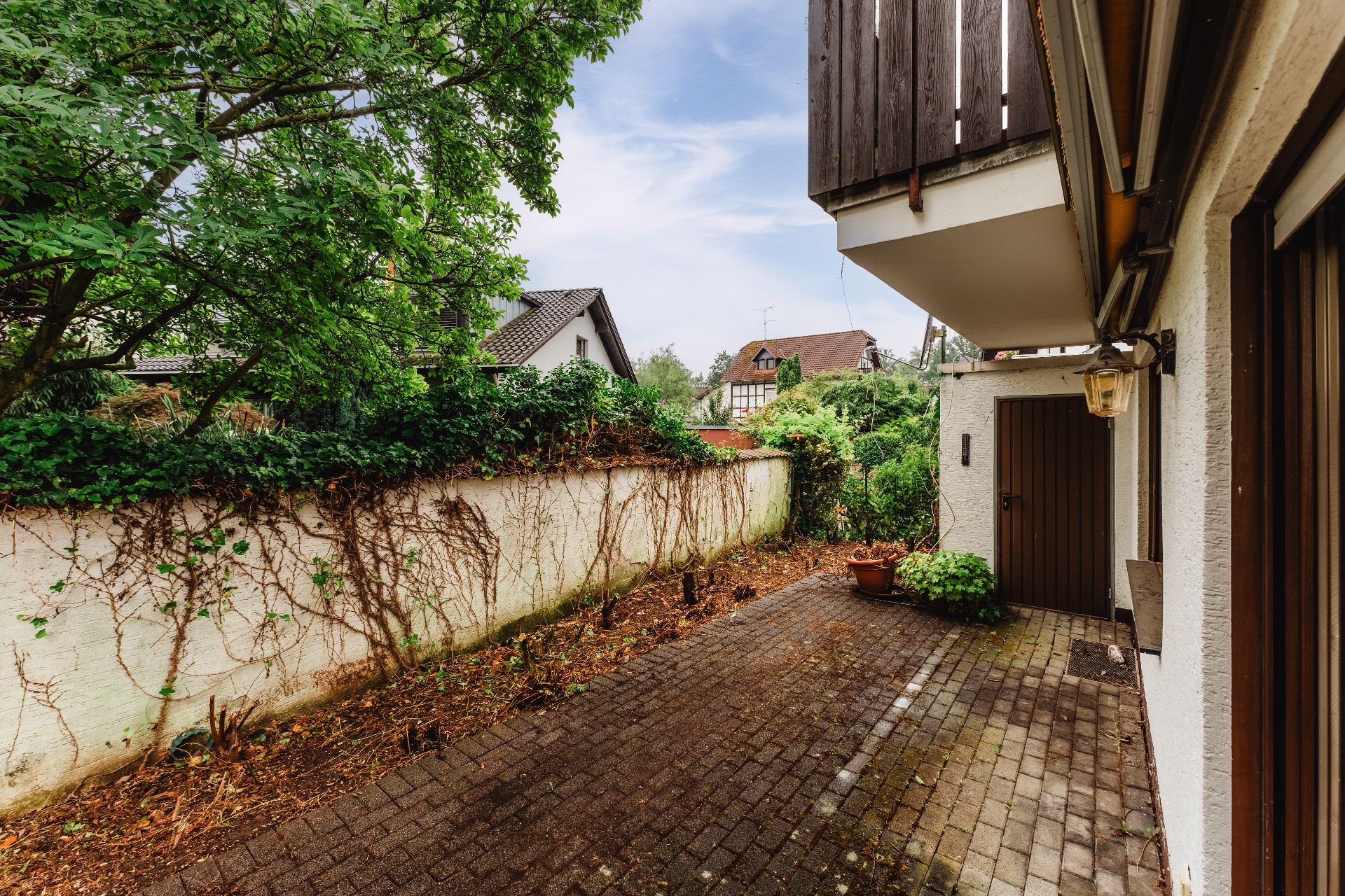 Gepflasterte Terrasse mit Topfpflanzen, Baum und angrenzender Mauer neben Wohnhaus.