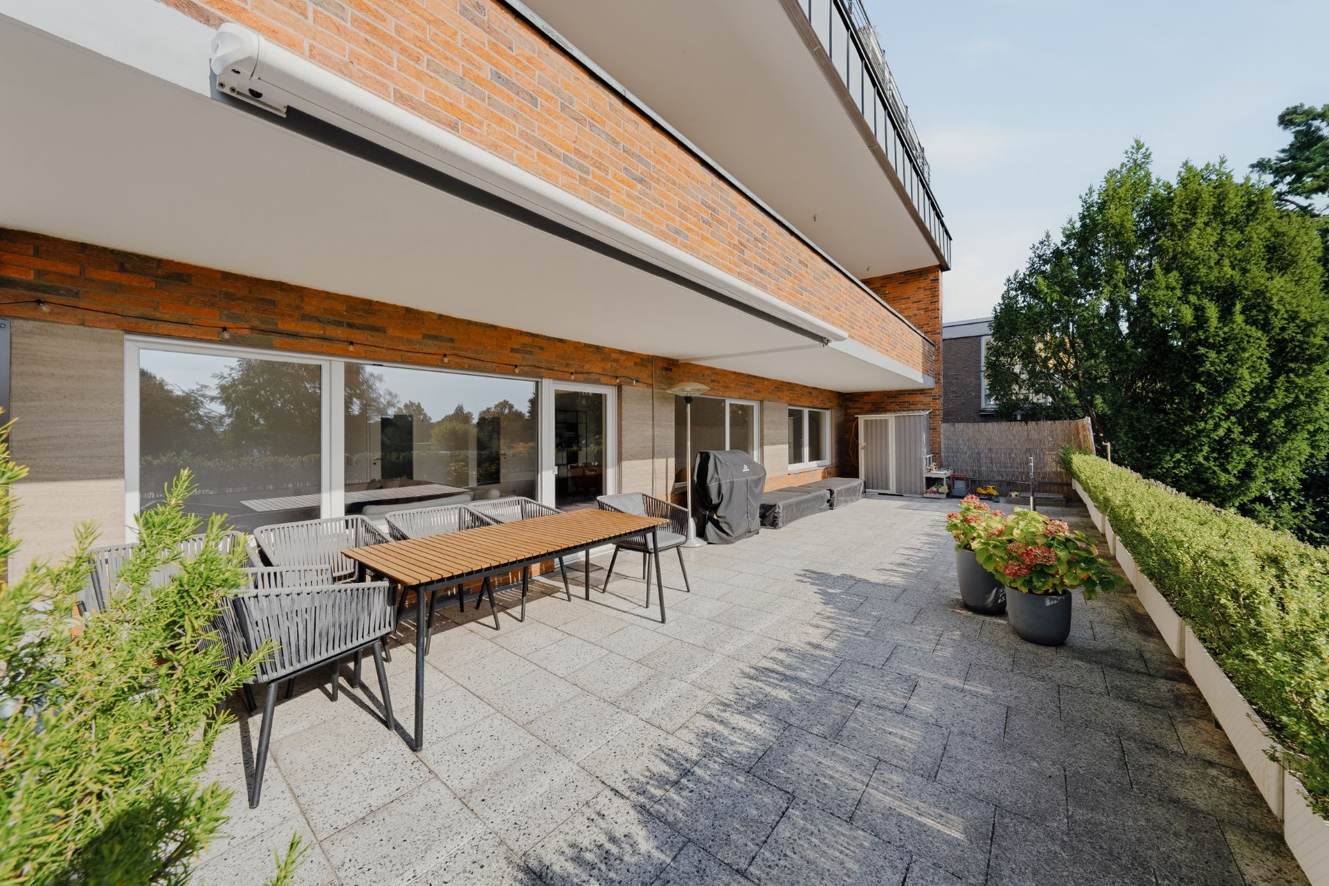 Große Terrasse mit Esstisch, Stühlen, Pflanzkübeln und Grill vor einem Wohnhaus.