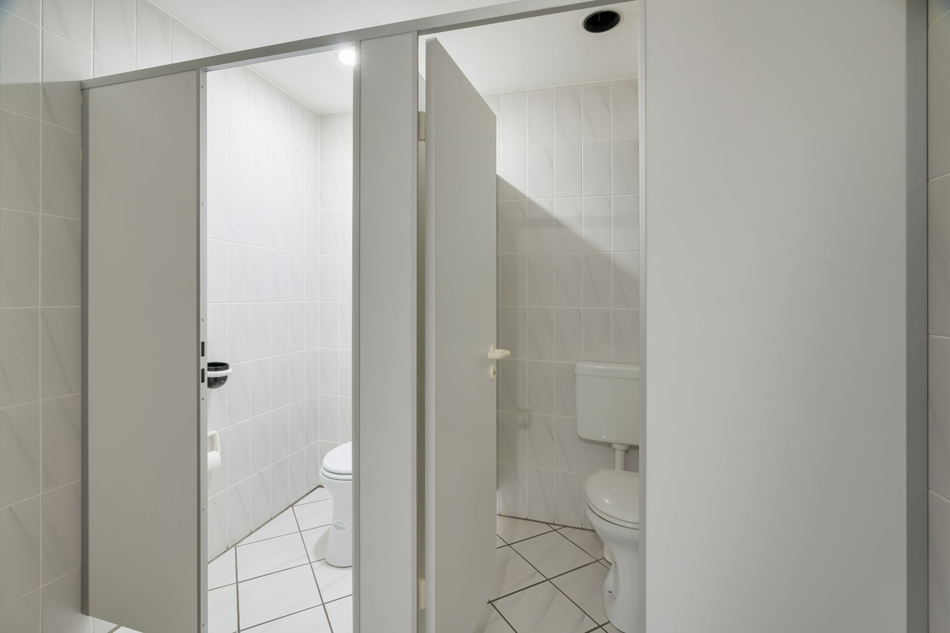 Zwei einzelne, weiße Toilettenkabinen mit Kacheln und offener Tür sind sichtbar.