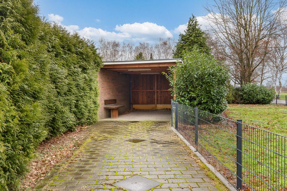 Gepflasterte Einfahrt mit Carport aus Holz, umgeben von Hecken und Zaun.