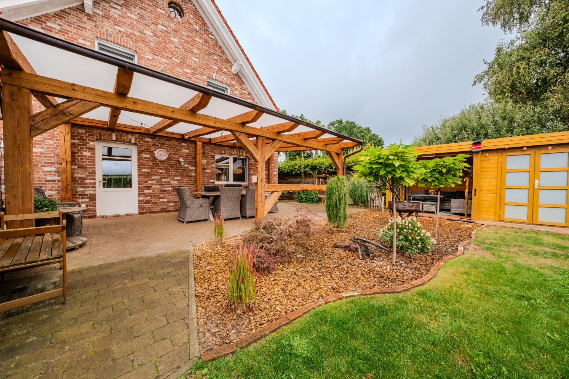 Überdachte Terrasse mit Sitzgruppe, Gartenbereich mit Bäumen und Holzhütte im Hintergrund.