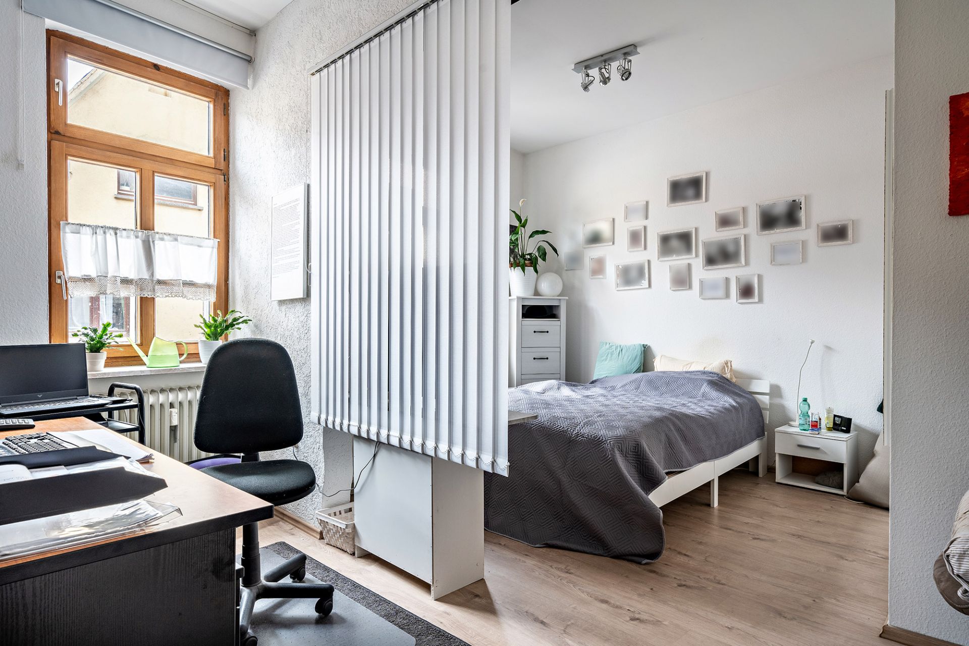 Helles Zimmer mit Schreibtisch, Stuhl und Bett, durch Lamellenvorhang getrennt.