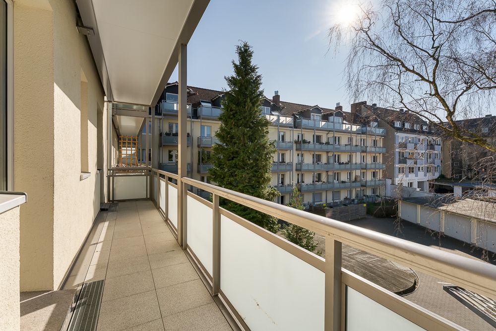 Langer Balkon mit Glasgeländer, Blick auf benachbarte Wohngebäude und Bäume bei Sonnenschein.