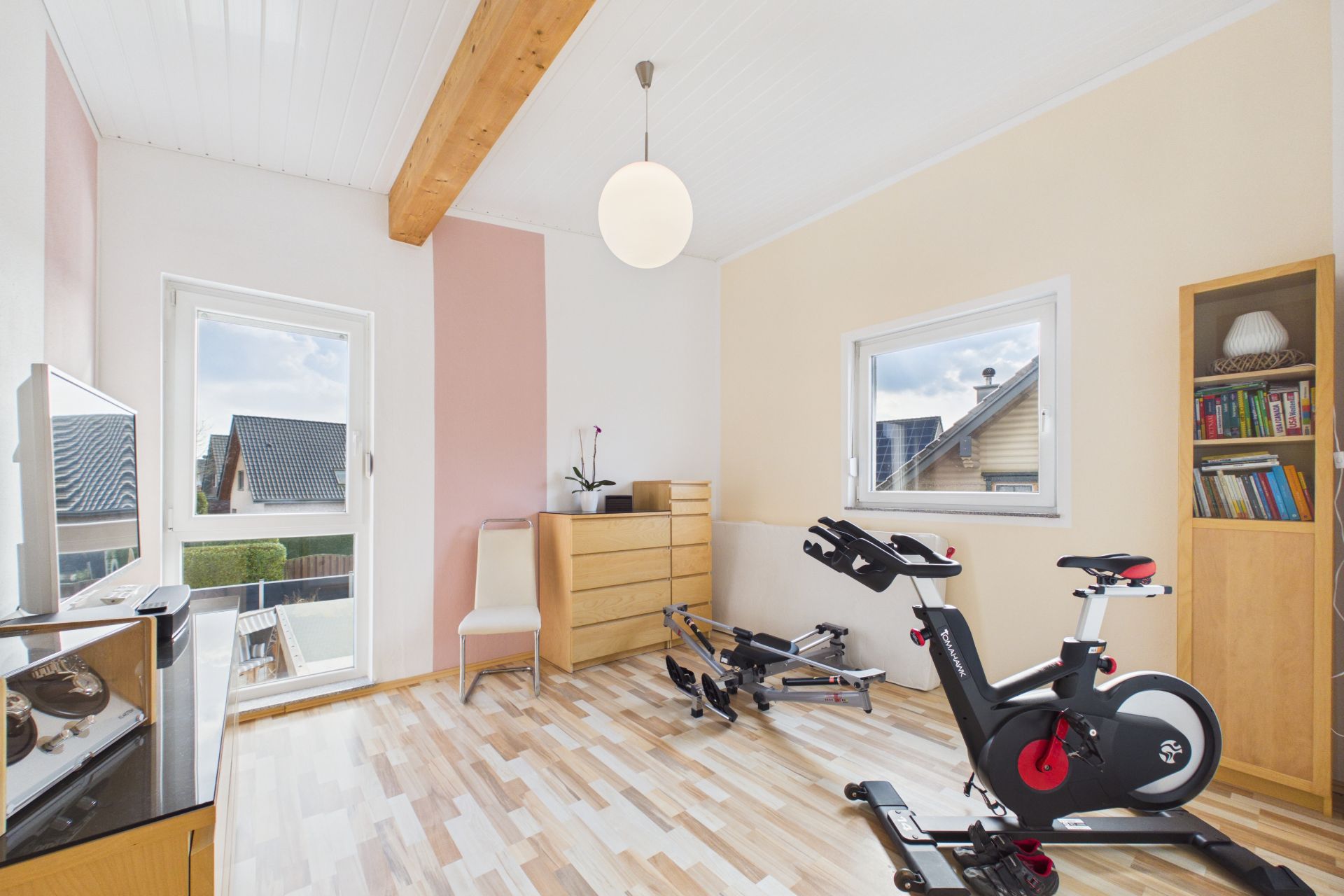 Heller Raum mit Fitnessgeräten, Regal, Kommode, Stuhl und großen Fenstern.
