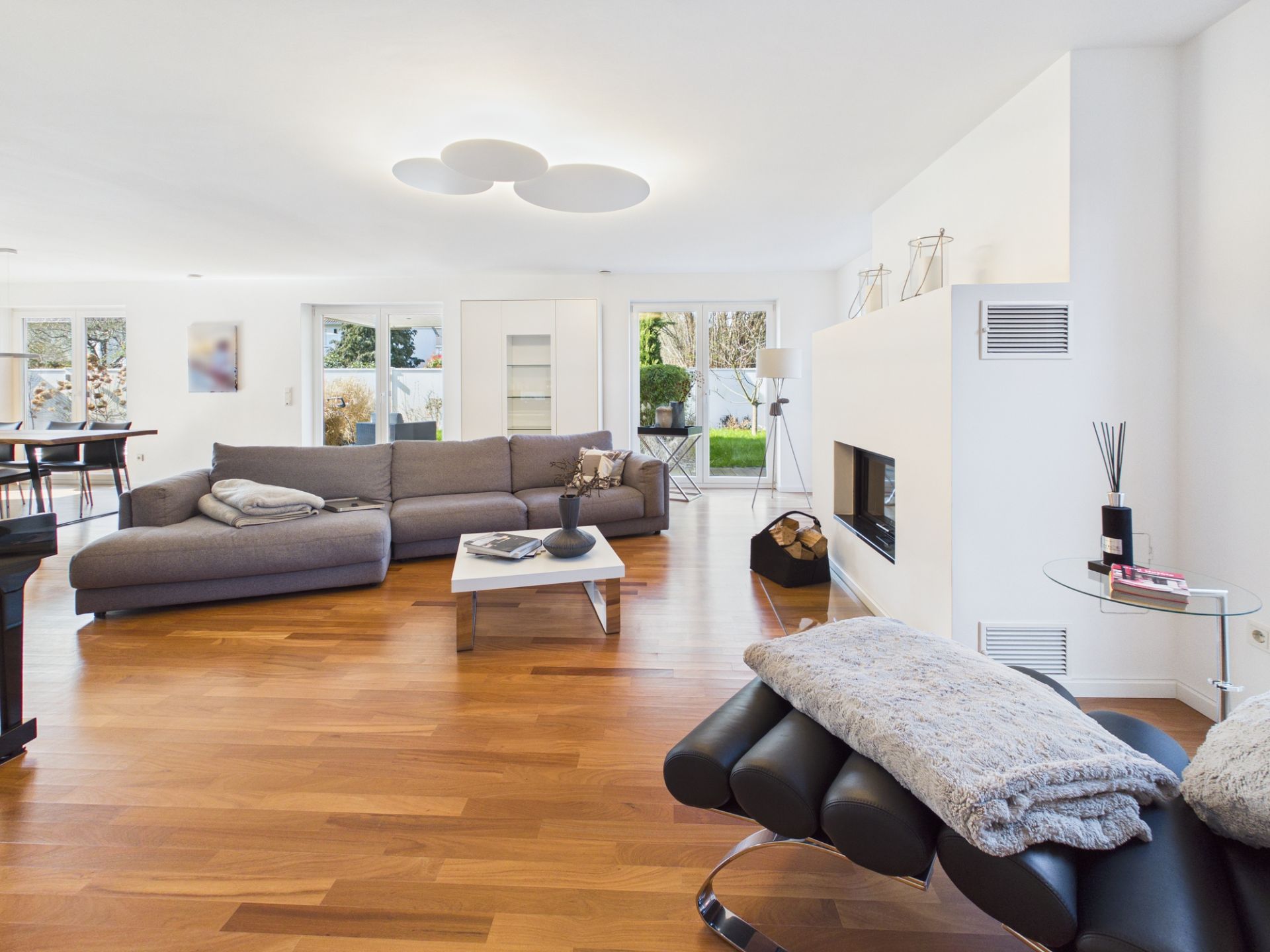 Modernes Wohnzimmer mit grauem Sofa, Kamin und Parkettboden, große Fenster bieten Blick in den Garten.