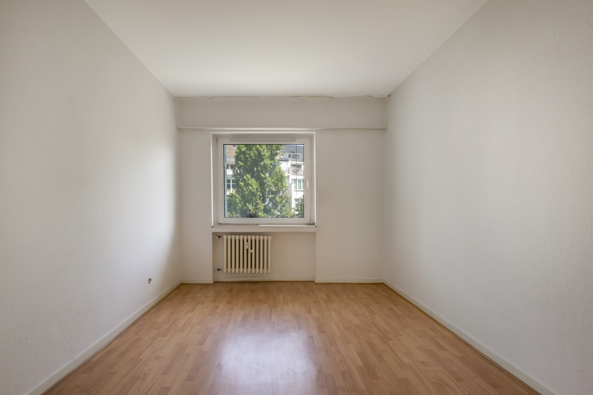 Leerer Raum mit Holzfußboden, weiß gestrichenen Wänden und einem Fenster mit Blick nach draußen.