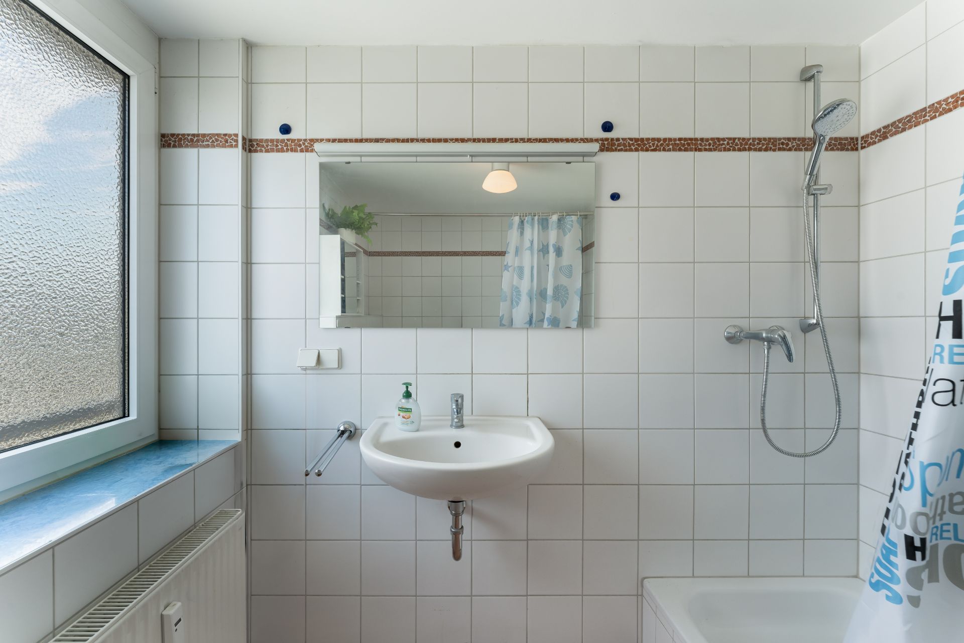 Weiß gefliestes Badezimmer mit Waschbecken, Spiegel, Dusche und Fenster.