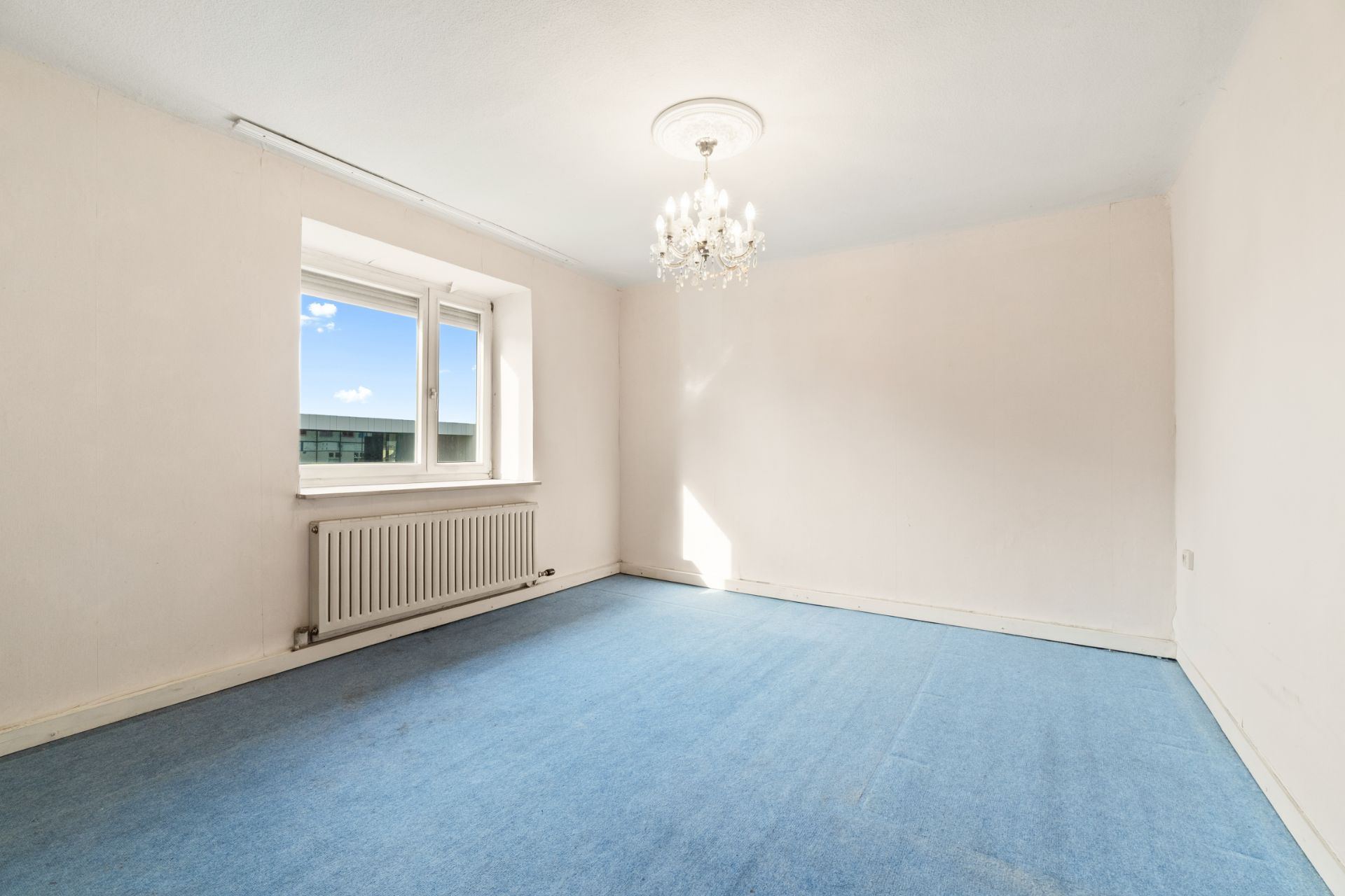 Helles, leeres Zimmer mit blauem Teppich, Fenster, Heizung und Kronleuchter an der Decke.