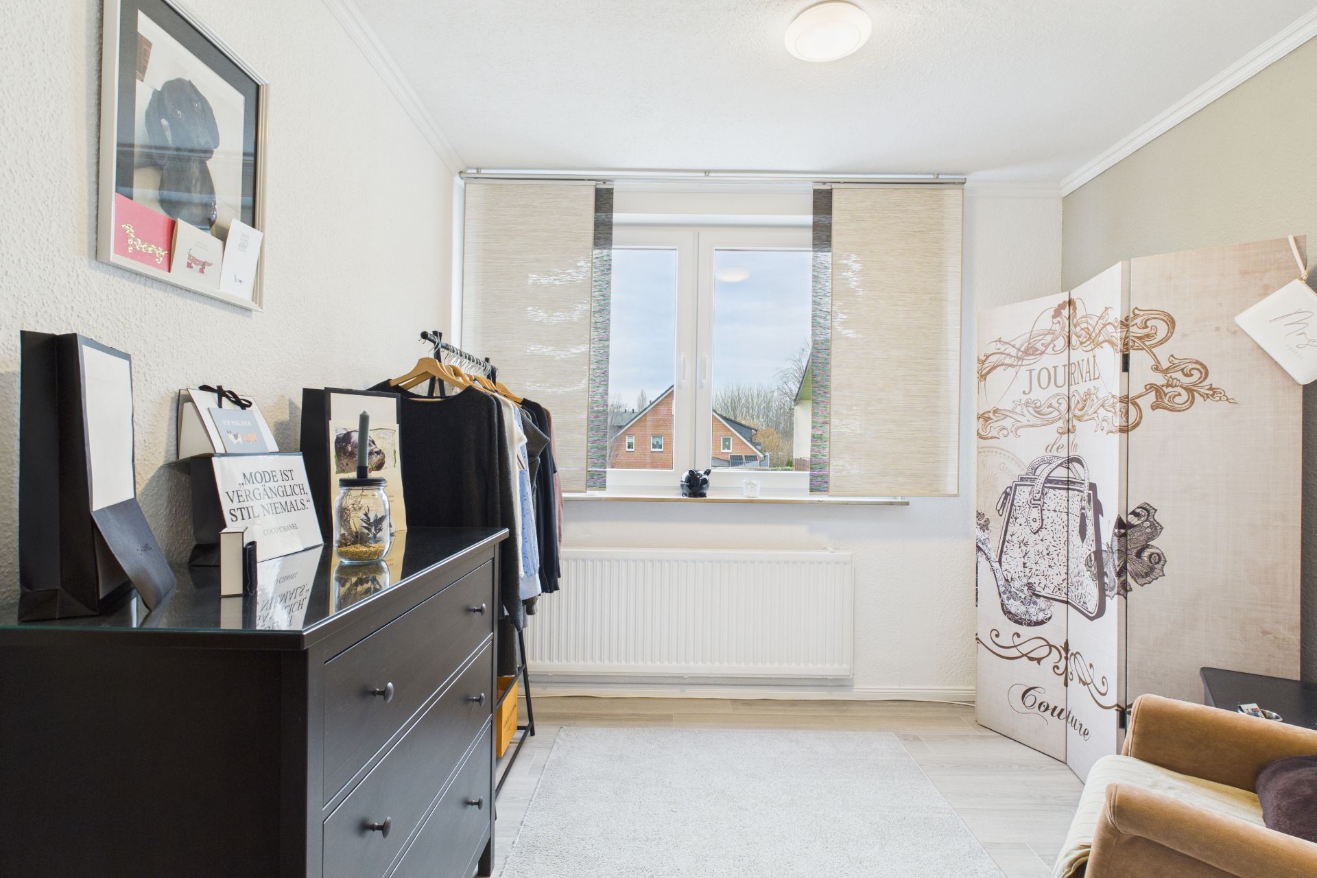 Helles Zimmer mit Kommode, Kleiderstange, Paravent und Fenster mit Blick nach draußen.