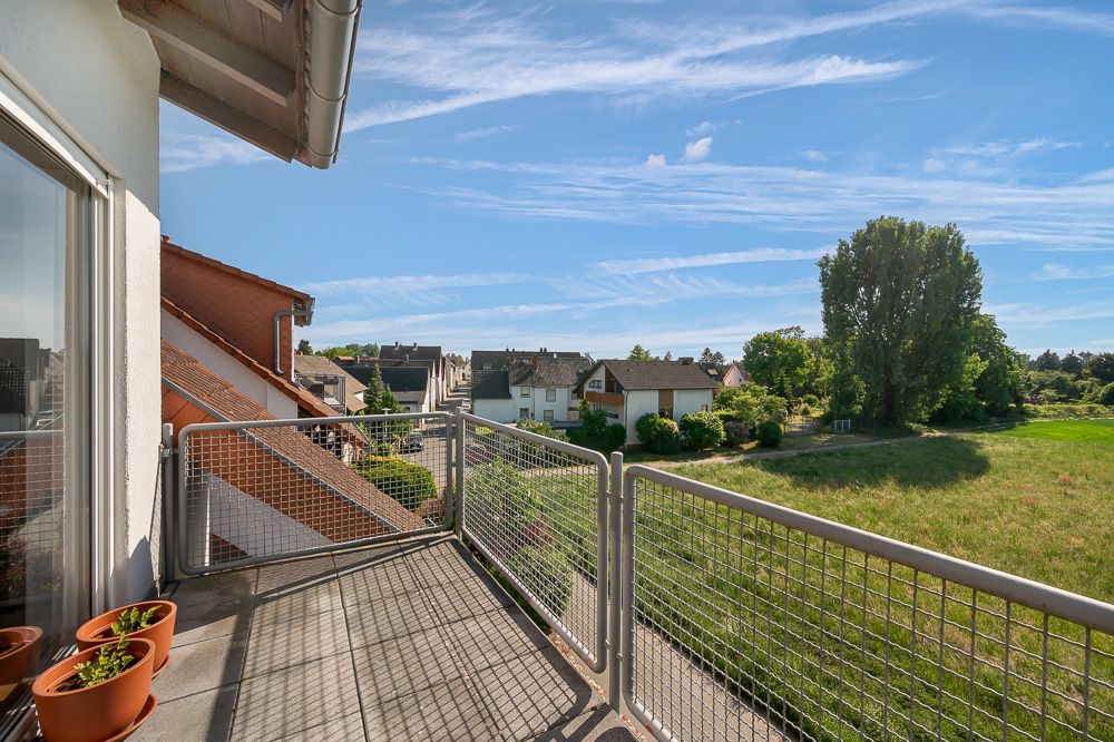 Balkon mit Metallgeländer, mehreren Blumentöpfen und Blick auf Wiese sowie Wohnhäuser.