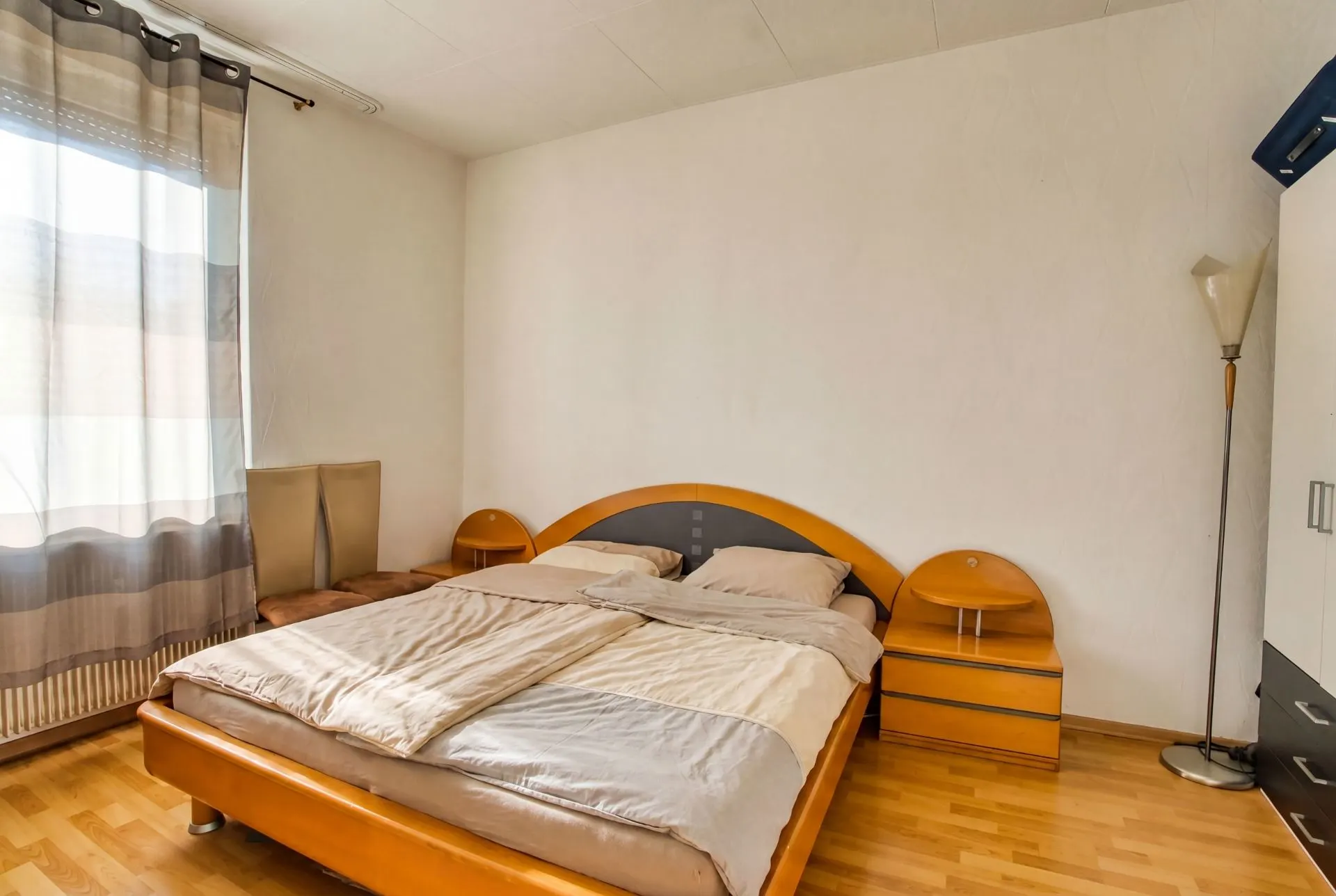 Schlafzimmer mit Doppelbett, zwei Nachttischen, Stehlampe und großem Fenster mit Vorhang.