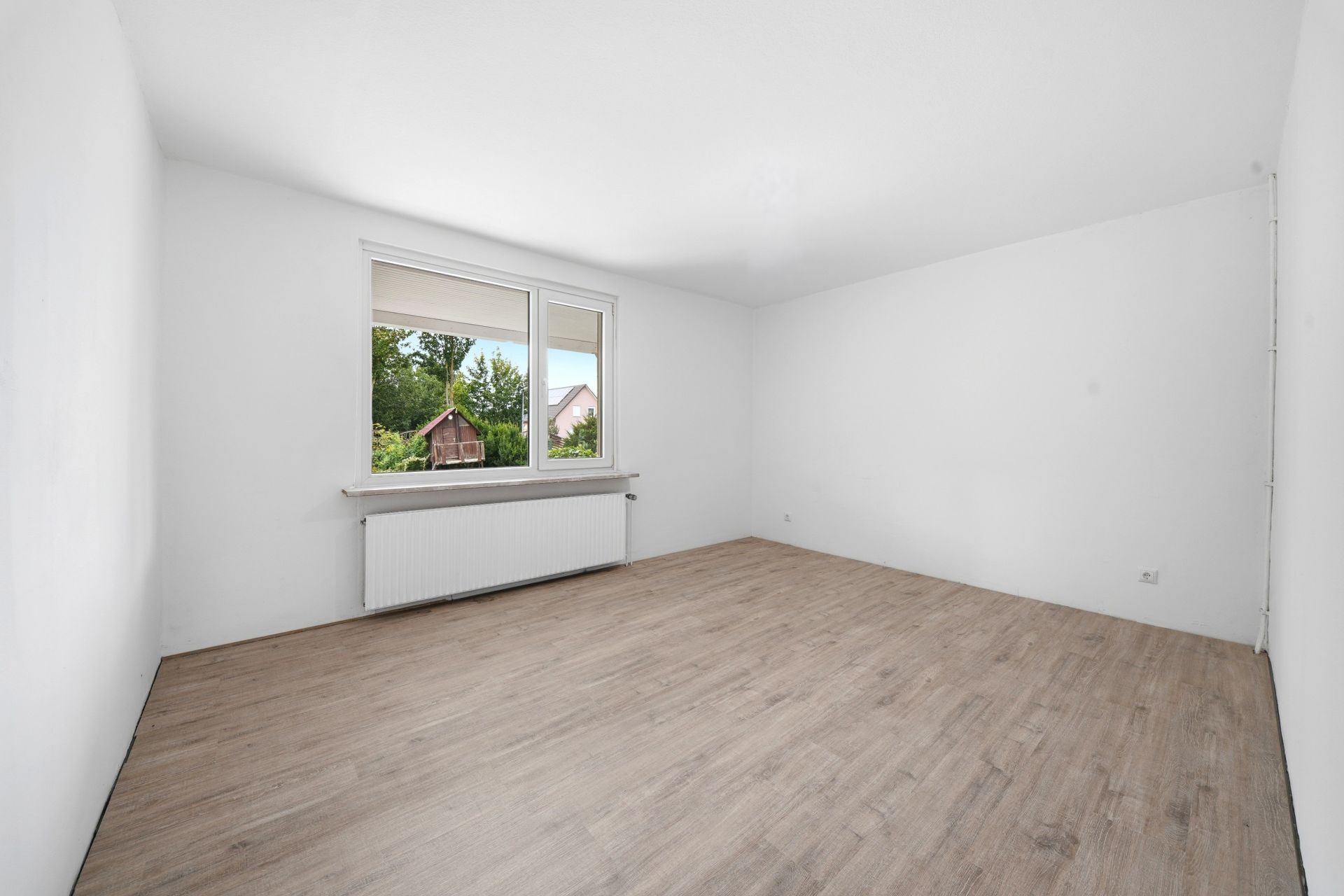 Leerer Raum mit hellem Holzfußboden, weißen Wänden und großem Fenster mit Blick ins Grüne.