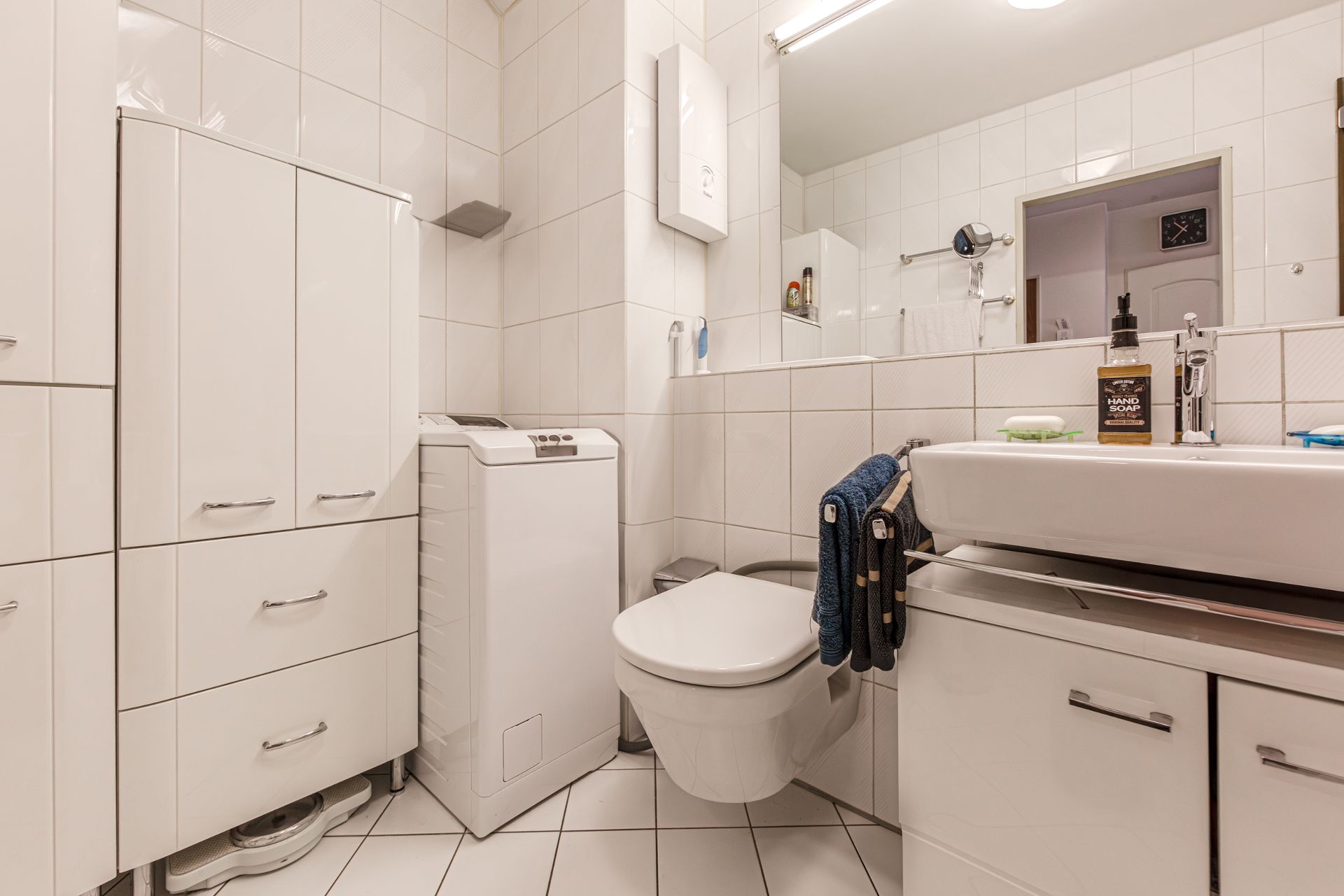 Helles Bad mit Waschmaschine, Hängetoilette, Waschbecken, Spiegel und Schränken.