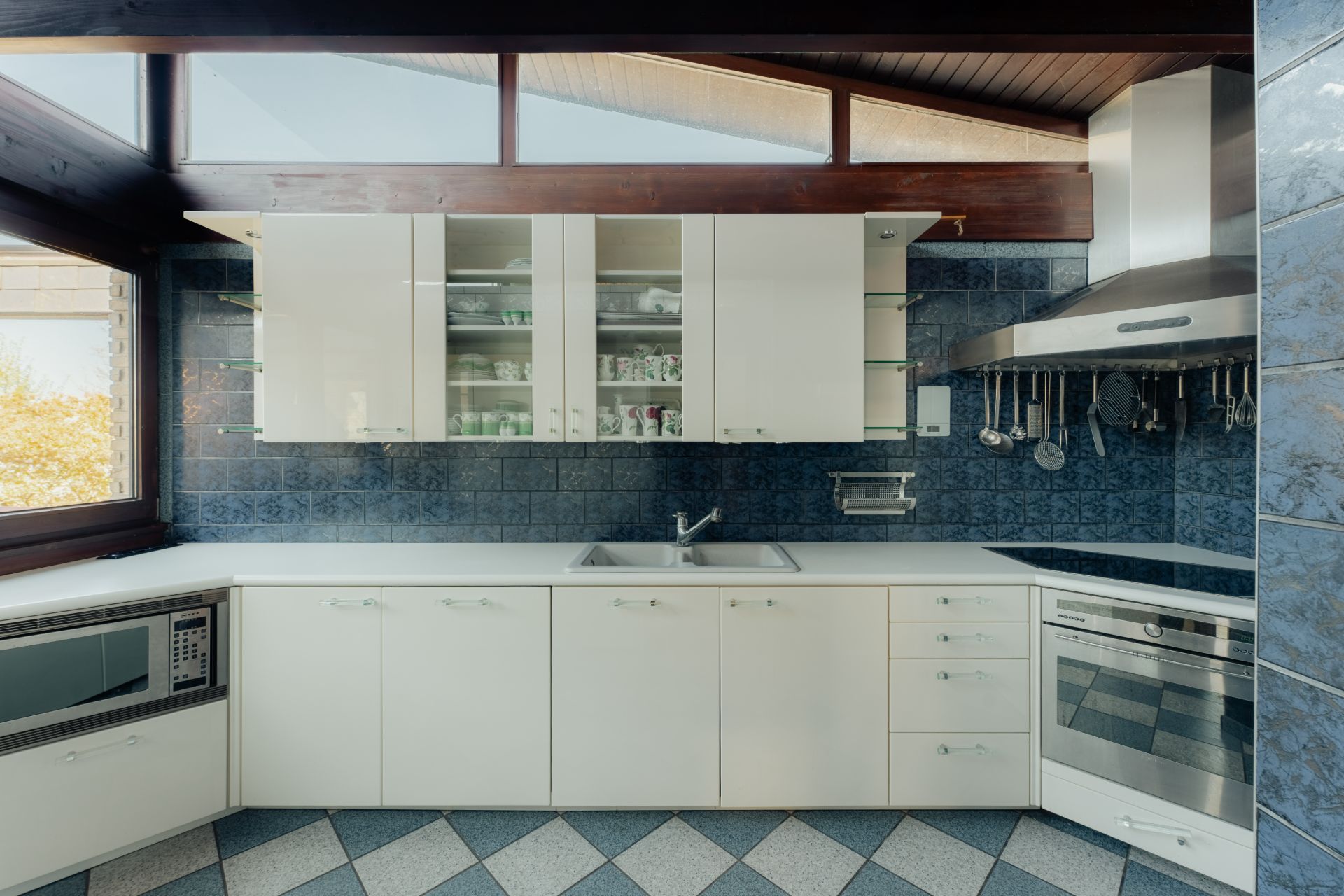 Moderne weiße Küche mit Hängeschränken, Edelstahlgeräten und blau gefliester Wand.