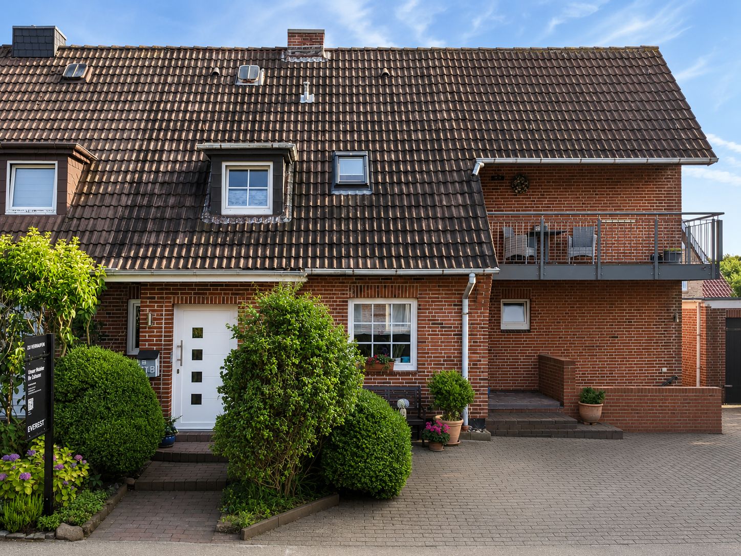 Ziegelhaus mit weißer Eingangstür, gepflegtem Vorgarten und Balkon auf der rechten Seite.