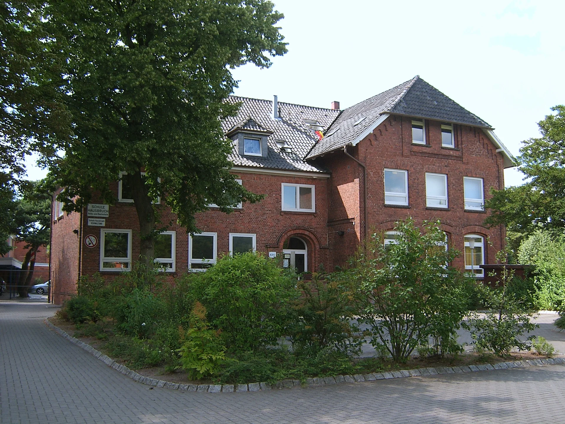 Backsteinhaus mit mehreren Fenstern, Bäumen und einem bepflanzten Vorgarten.