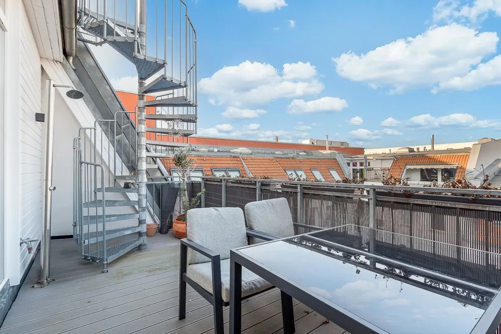 Möblierte Terrasse mit Tisch, zwei Stühlen und Wendeltreppe, umgeben von Häuserdächern.